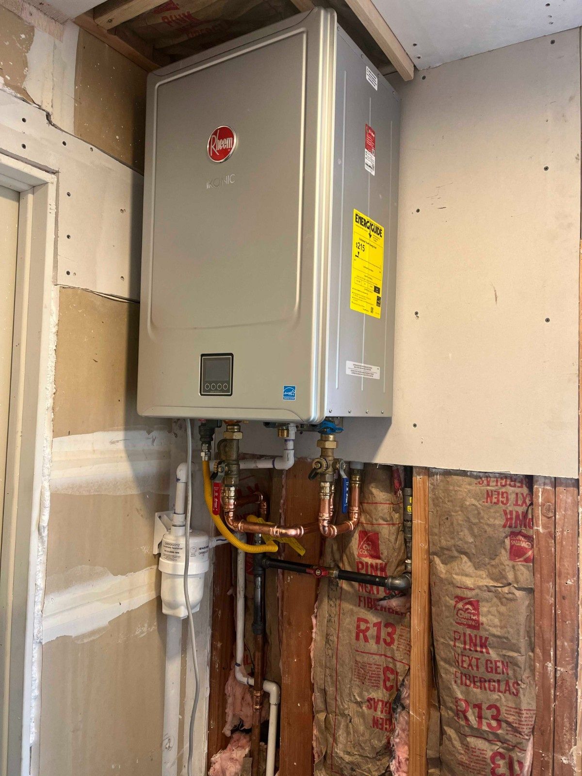 A silver Rheem tankless water heater mounted on an unfinished wall with exposed insulation and plumbing connections.