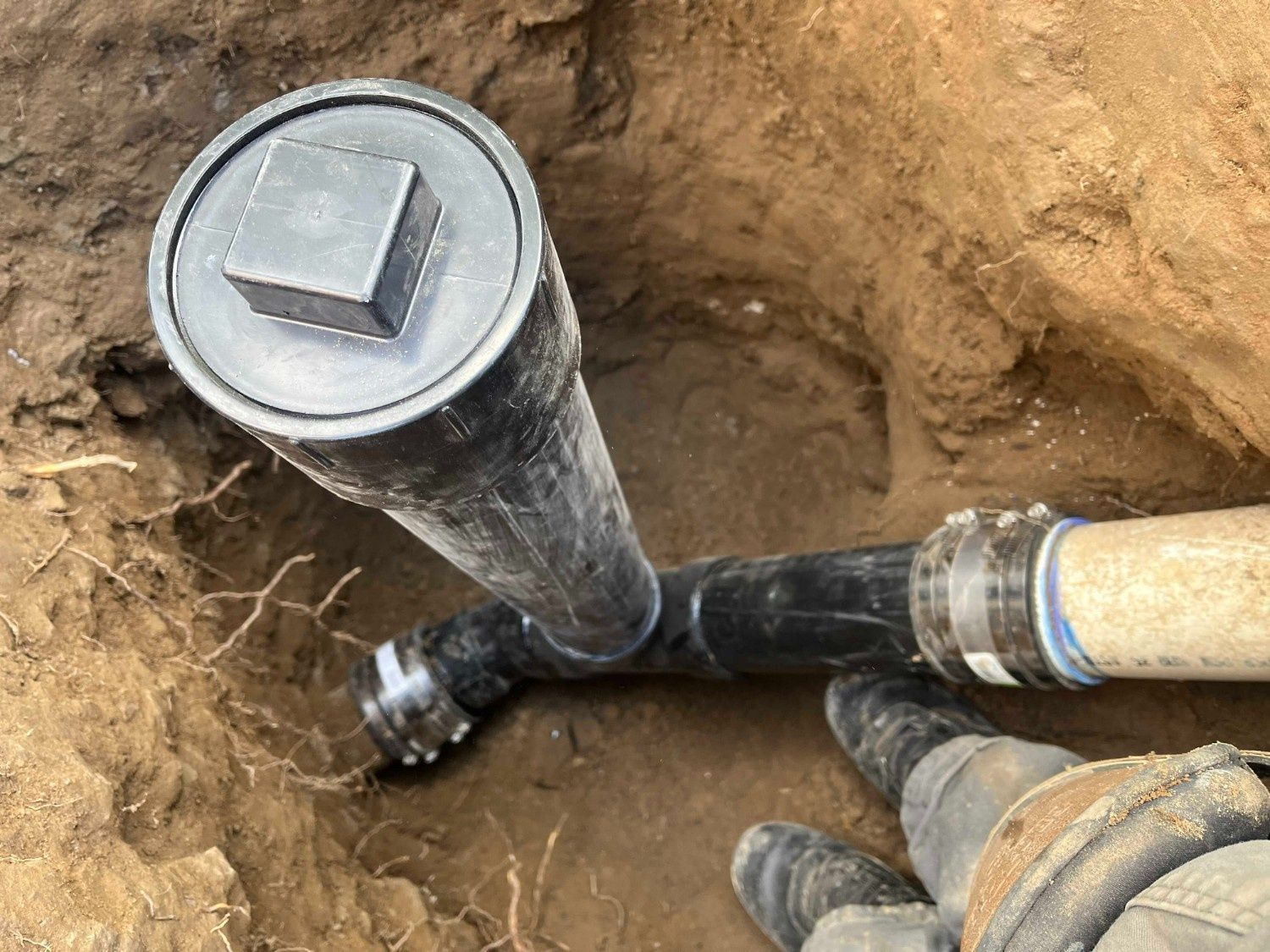 A close-up view of a black plastic sewer cleanout pipe installed in a dirt trench with a rubber coupling connector.
