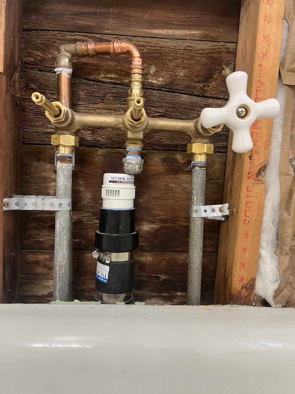 Exposed plumbing fixtures and copper pipes attached to wooden wall studs behind a bathtub.