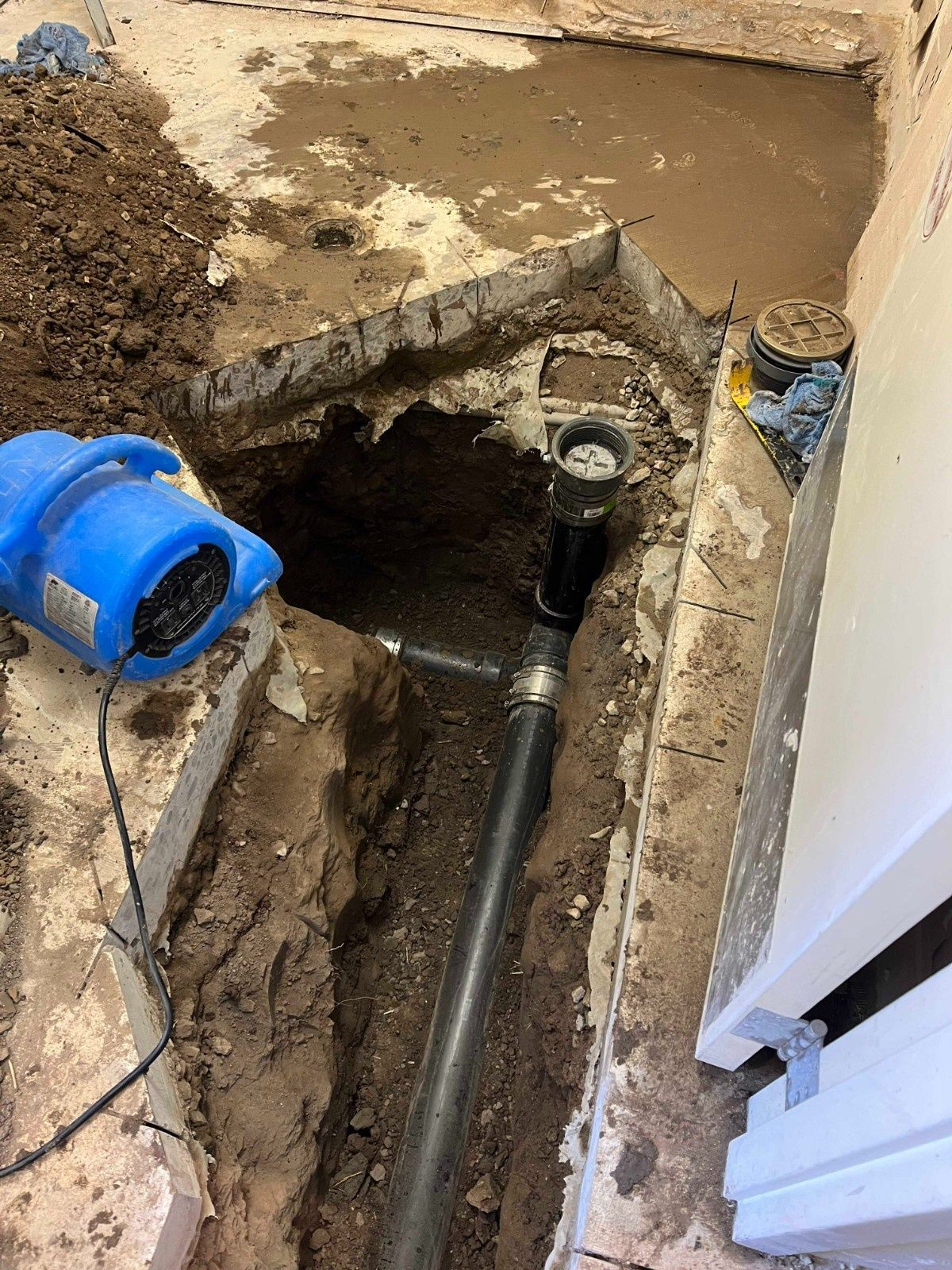 A trench dug in a concrete floor reveals a plumbing pipe with a cleanout cap, located next to a blue air mover.