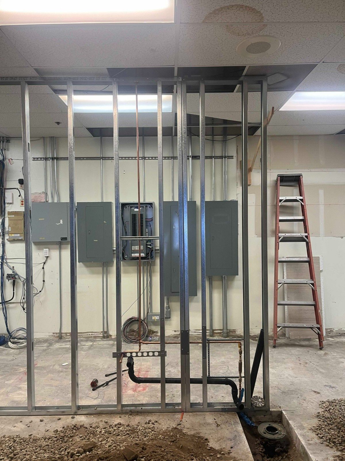Metal wall framing studs are erected in front of electrical panels and plumbing pipes inside a construction space.