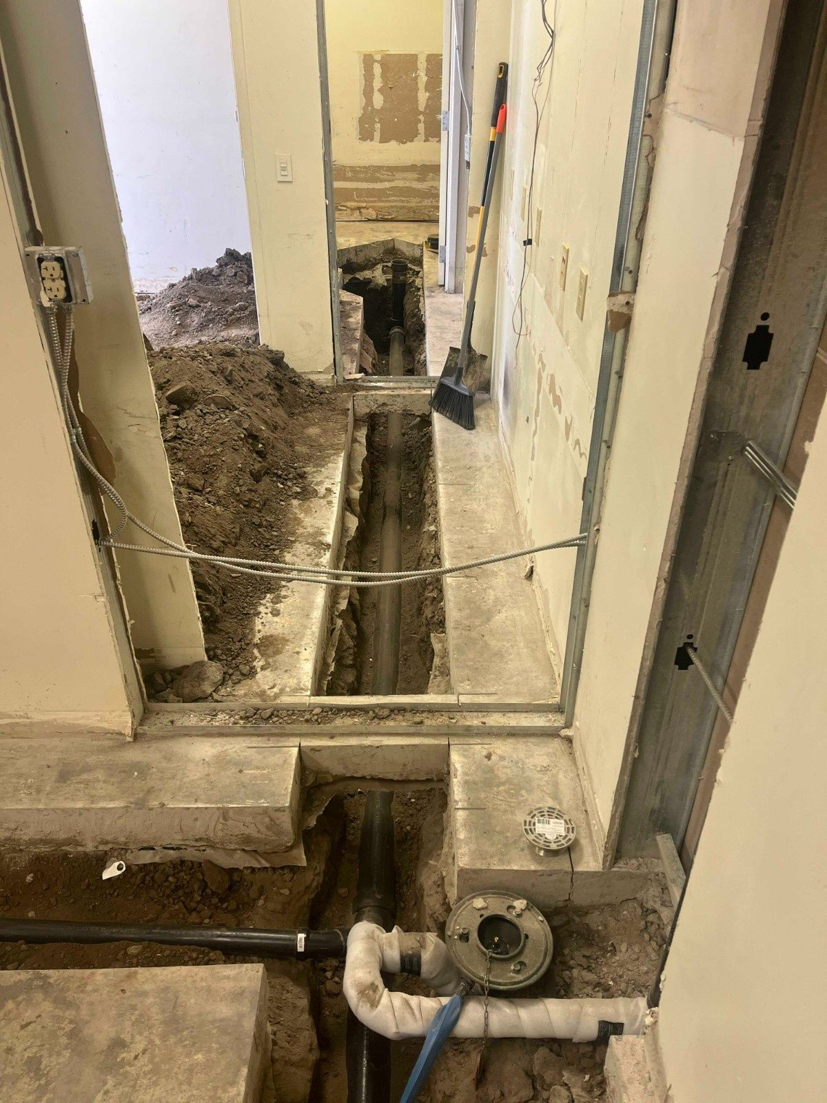 A trench cut through a concrete floor in a construction area, exposing dirt and plumbing pipes.