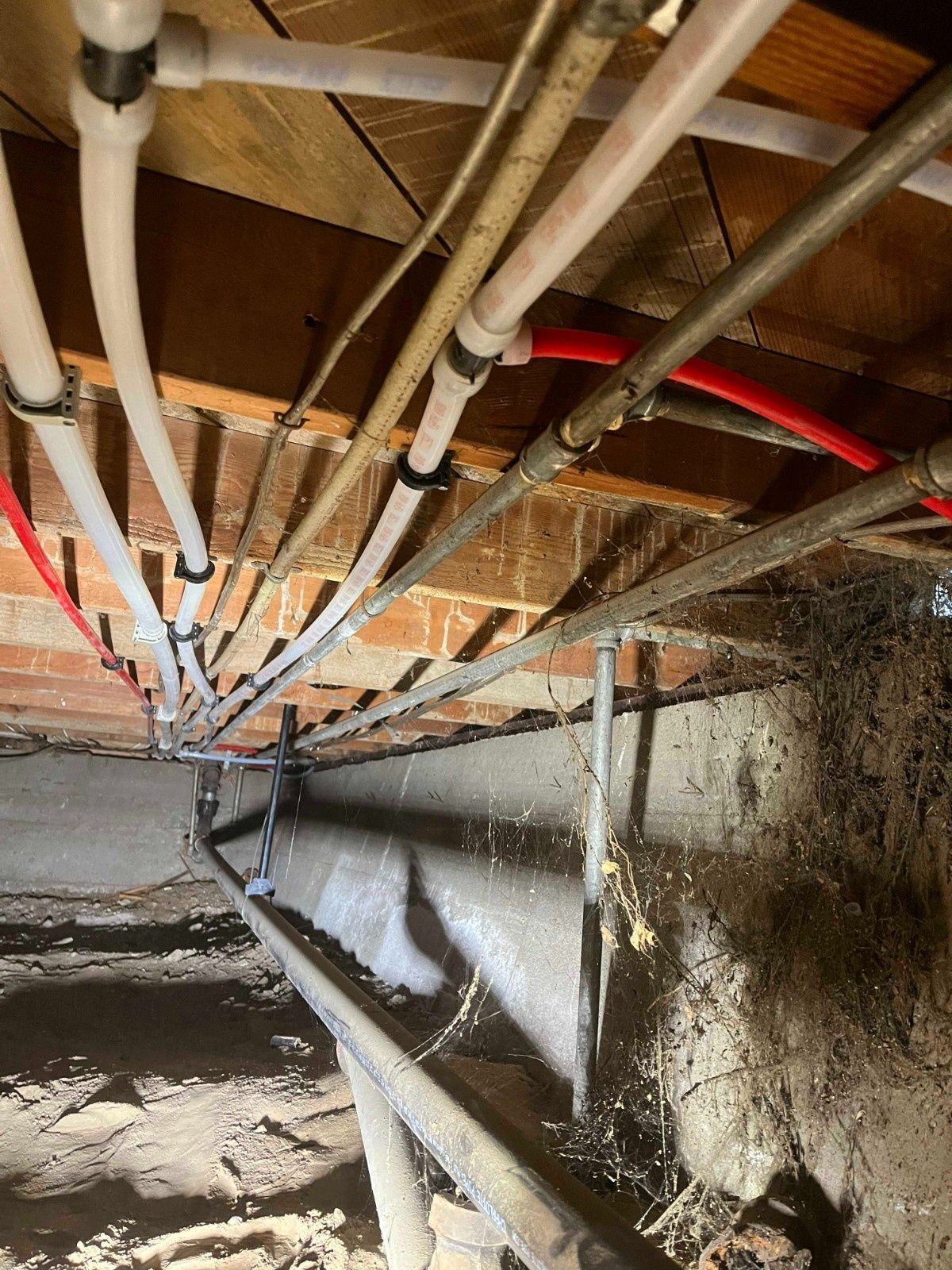 Crawl space showing PEX plumbing pipes mounted to wooden floor joists above a dirt floor and concrete foundation.