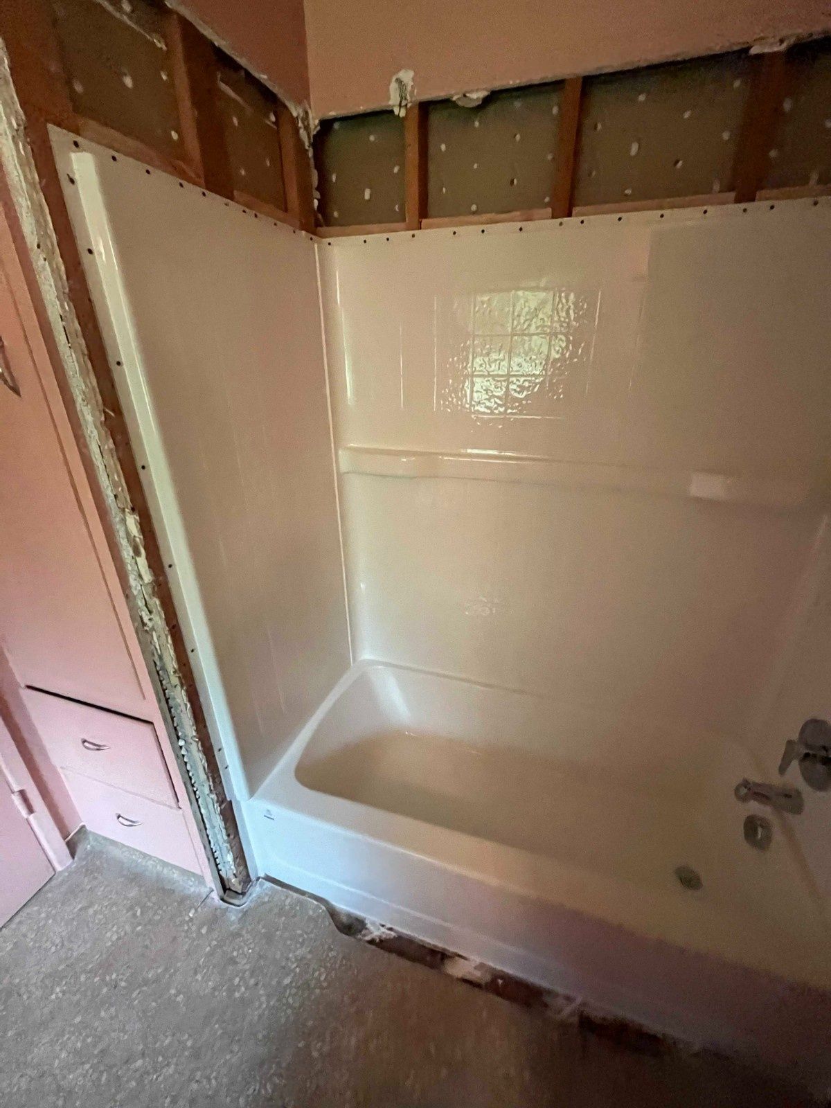 An unfinished bathroom showing a white bathtub and shower surround, with exposed wall studs and pink paint nearby.