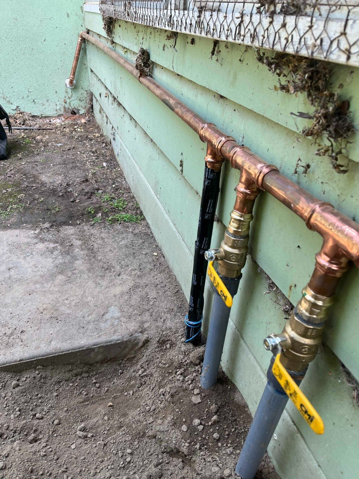 A copper plumbing manifold with two yellow-handled shut-off valves connected to grey piping along an exterior wall.