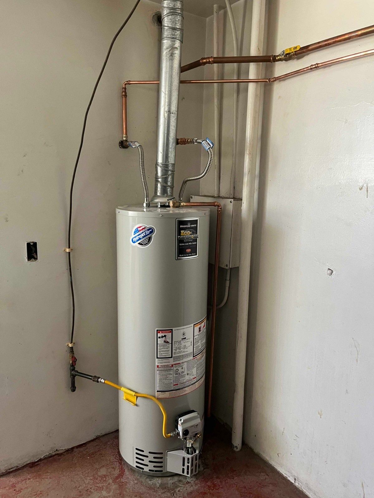 A gas water heater standing in the corner of a room, connected to copper piping and a metal exhaust vent.