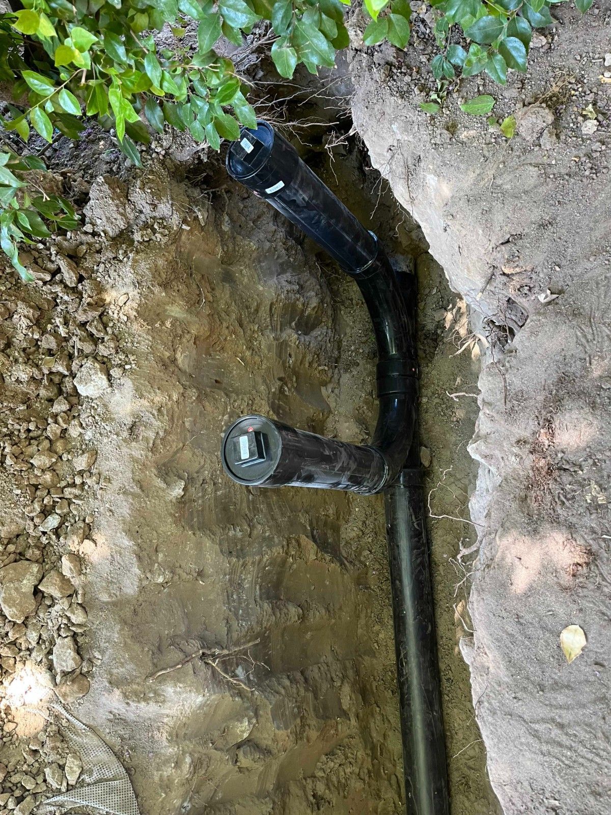 An exposed black pipe irrigation system with a valve control box set in a dirt trench outdoors.
