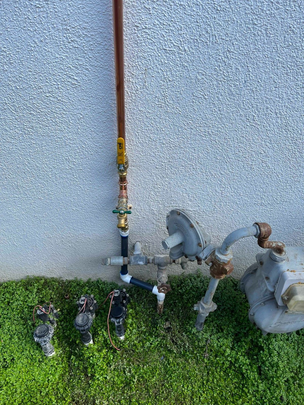 A gas meter assembly with pipes, valves, and a regulator mounted against a textured exterior wall over a grassy lawn.