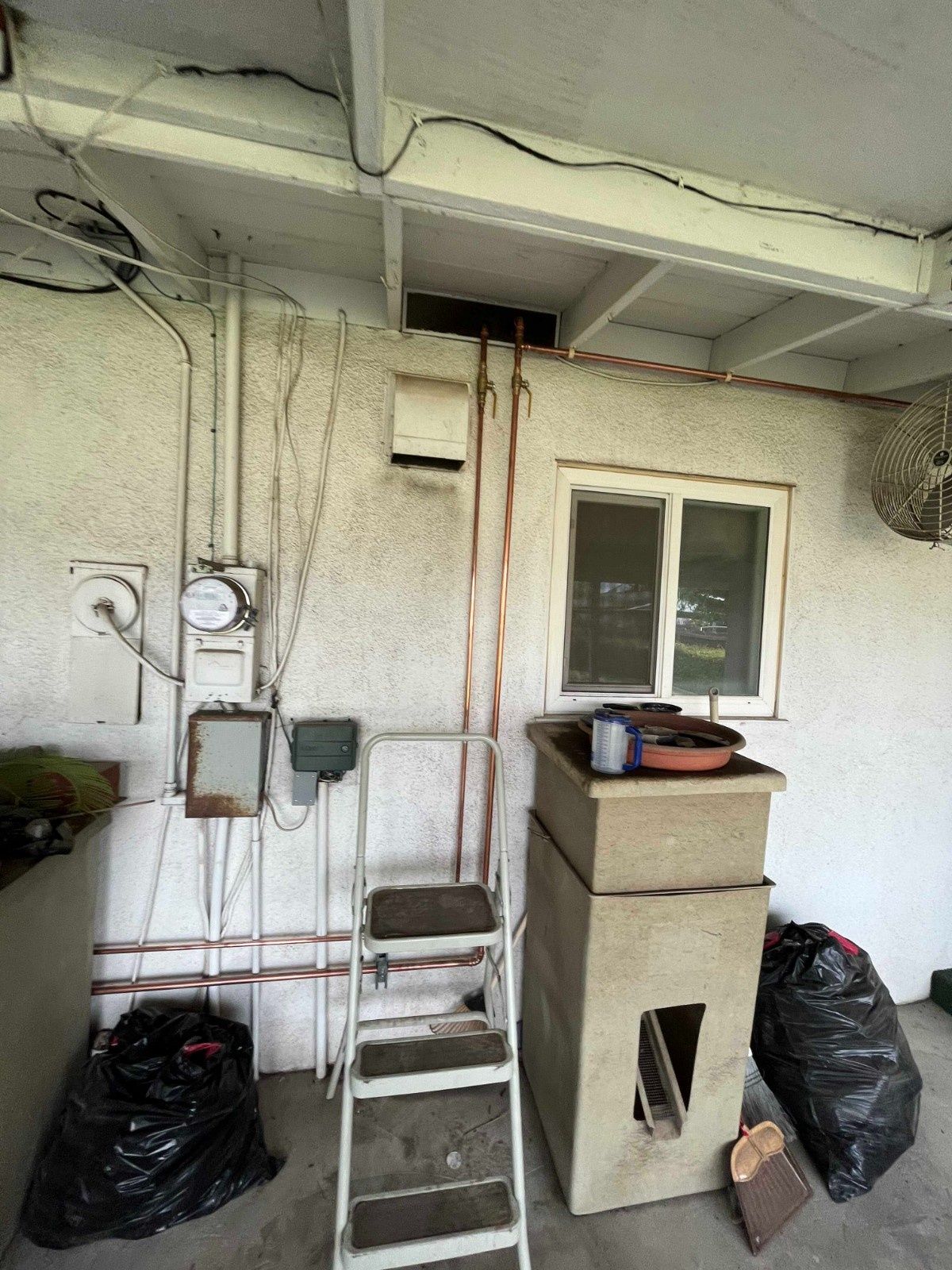 An outdoor utility area with exposed electrical wiring, copper pipes, a step ladder, a utility sink, and trash bags.