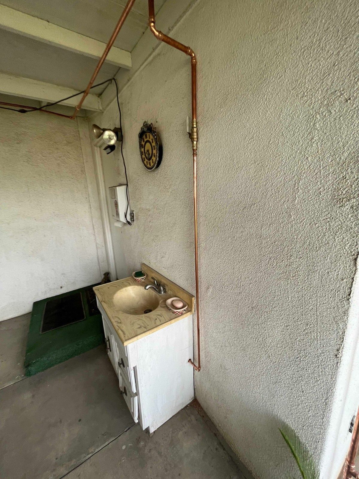 A small outdoor sink with a copper pipe running up the stucco wall, next to a dartboard on a covered patio.