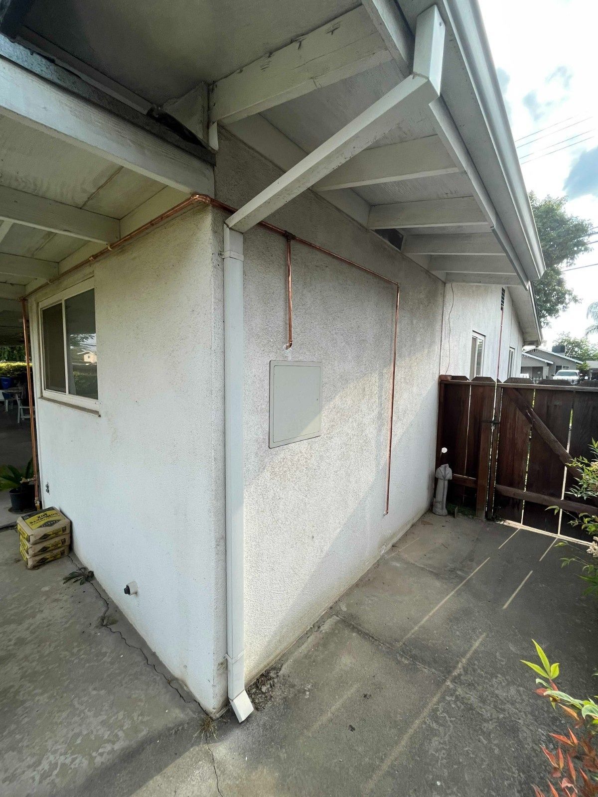 Exterior corner of a white stucco house with a downspout, an electrical panel, and a concrete patio.