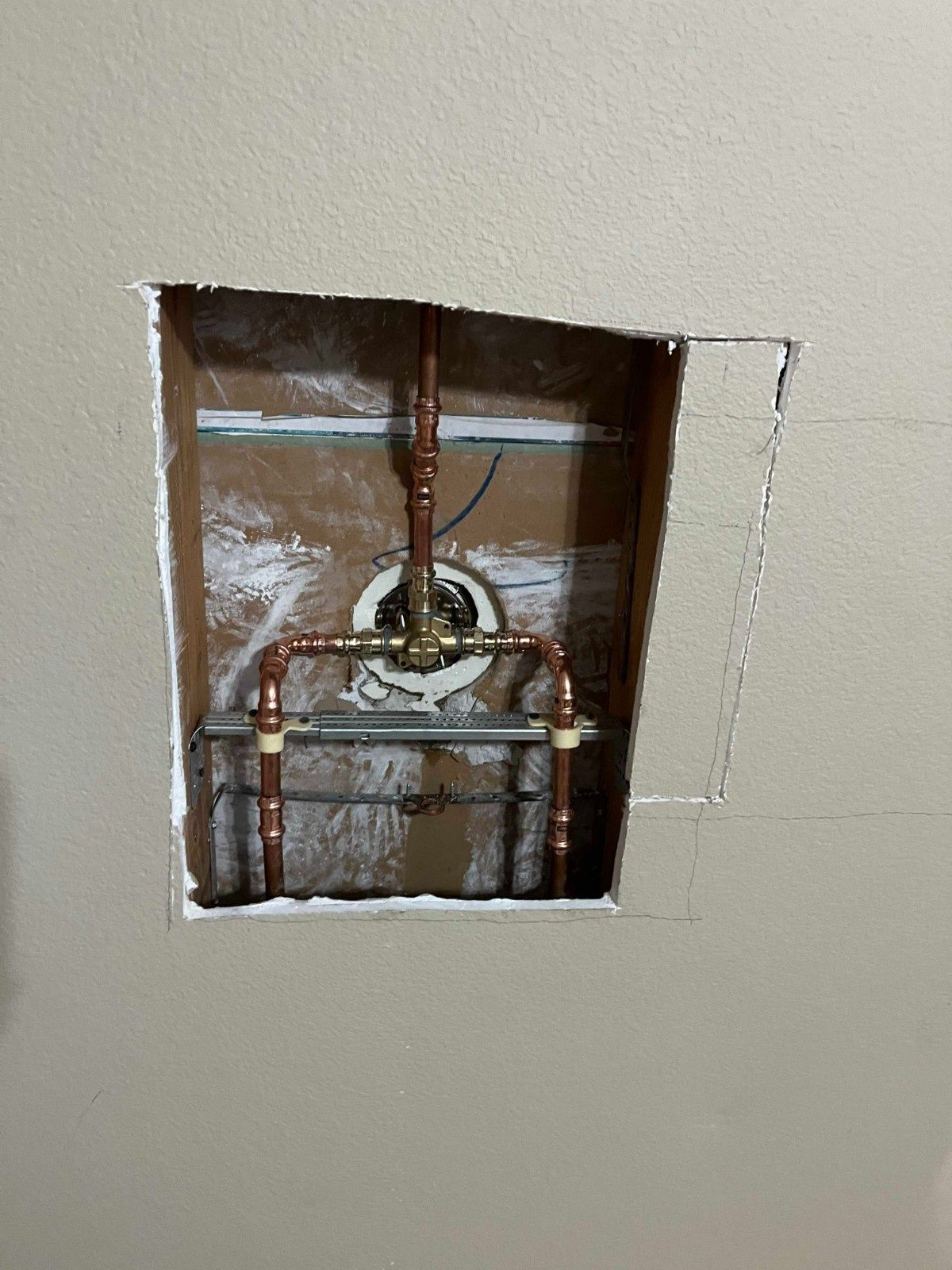 Copper plumbing pipes and a shower valve installed within an open rectangular hole in a beige drywall wall.