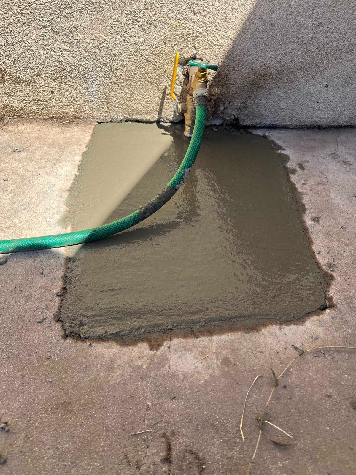 A patch of wet, gray cement fills a rectangular hole in a concrete ground surface, with a green garden hose lying across it.