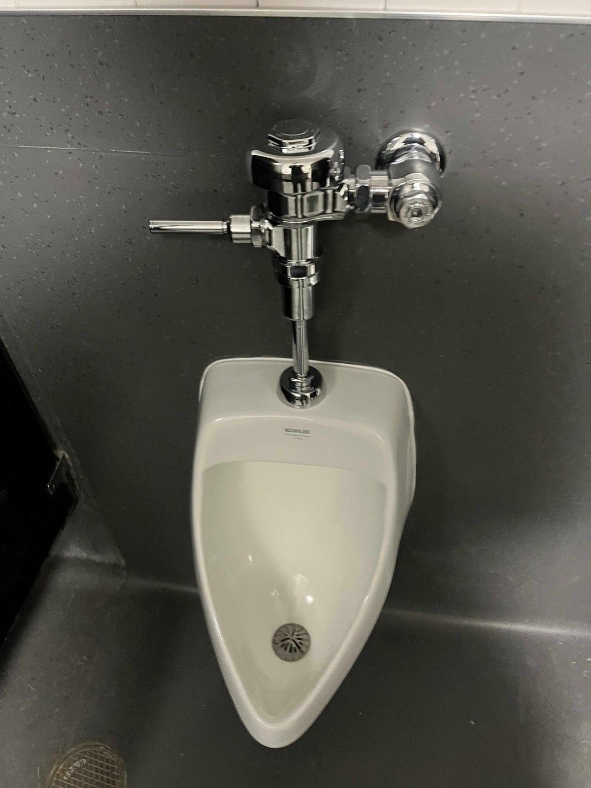 A wall-mounted white urinal with a chrome manual flush valve handle, centered on a dark grey partition wall.