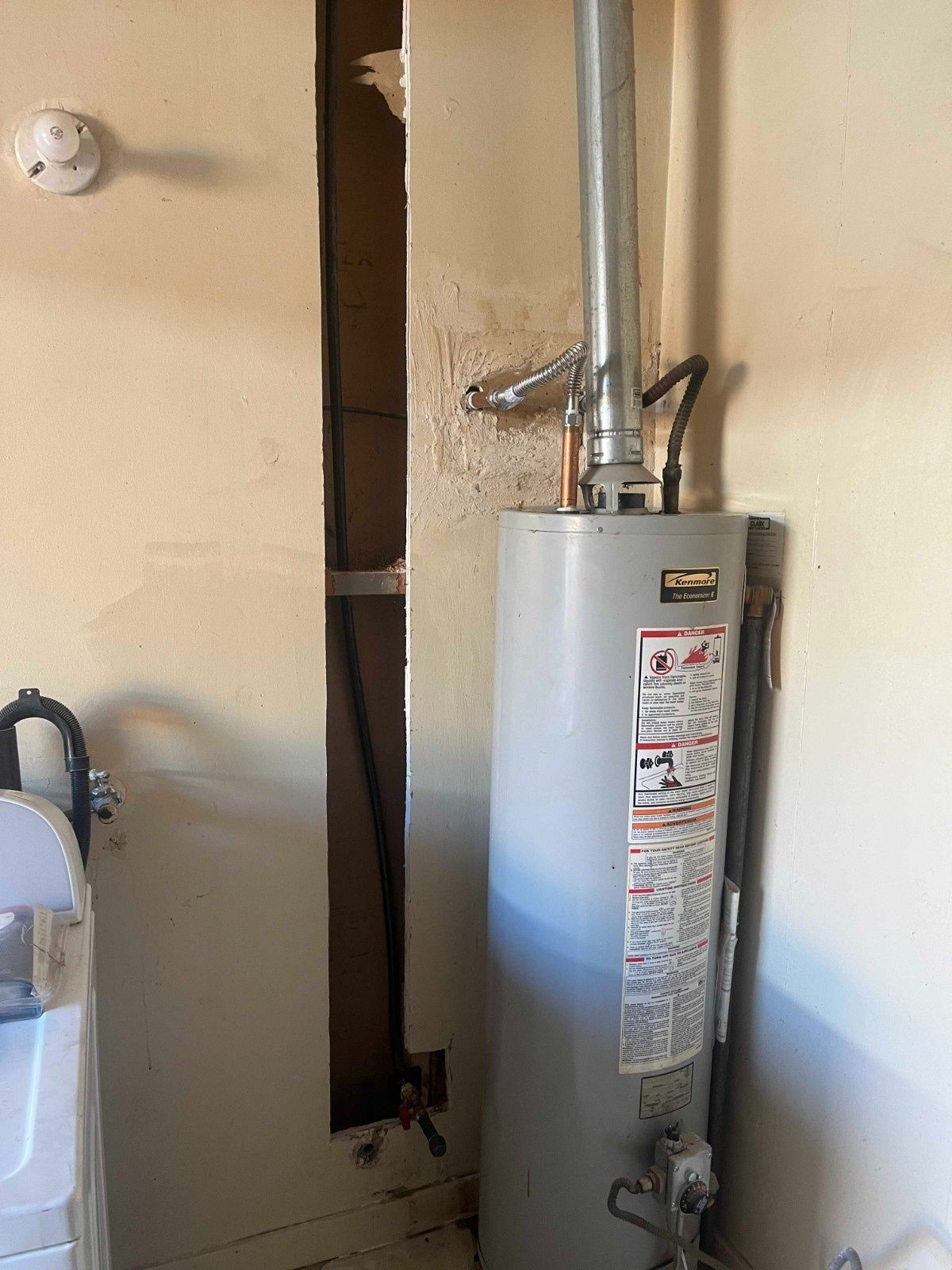 A grey water heater in a room with a vertical gap cut into the beige wall showing internal wiring and a pipe.