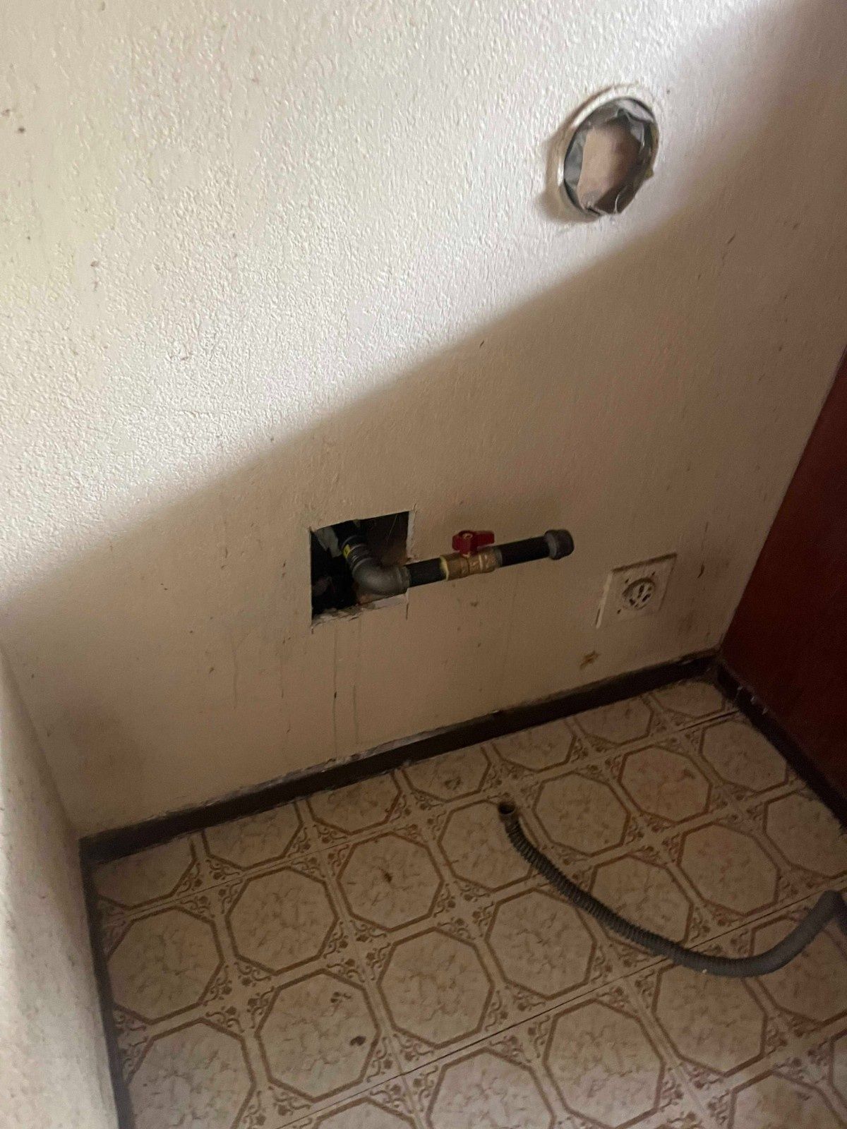 A gas line with a red shut-off valve protruding from a hole in a beige wall above a tiled floor.