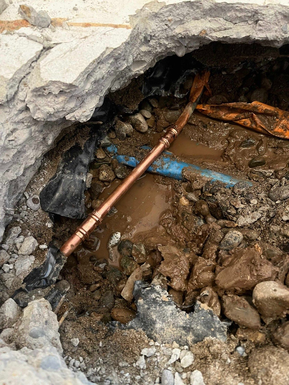 A copper pipe section repaired with couplings lies in a muddy, excavated hole through concrete pavement.