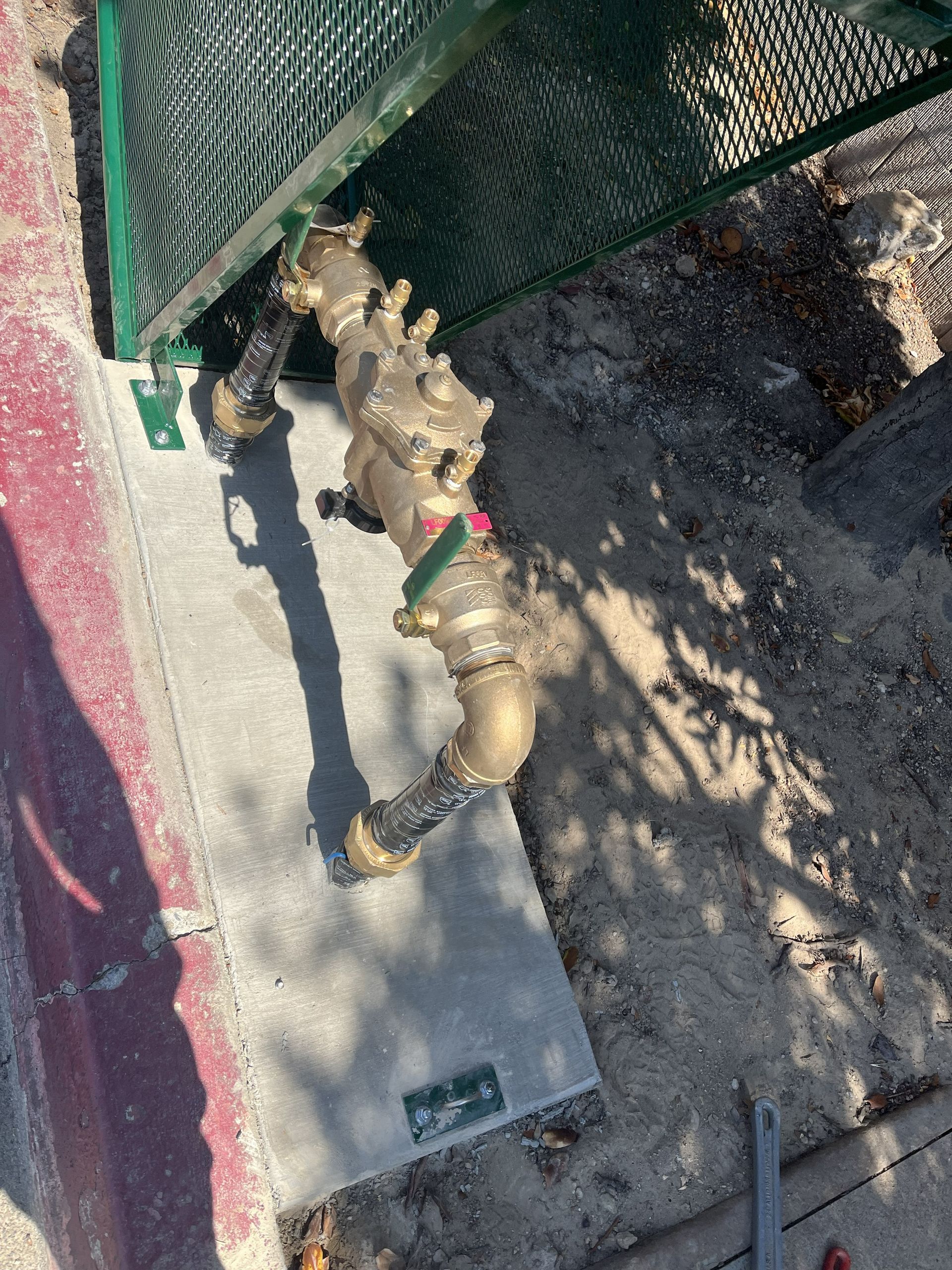 A bronze-colored backflow prevention device installed on a concrete pad next to a curb and a green mesh screen.