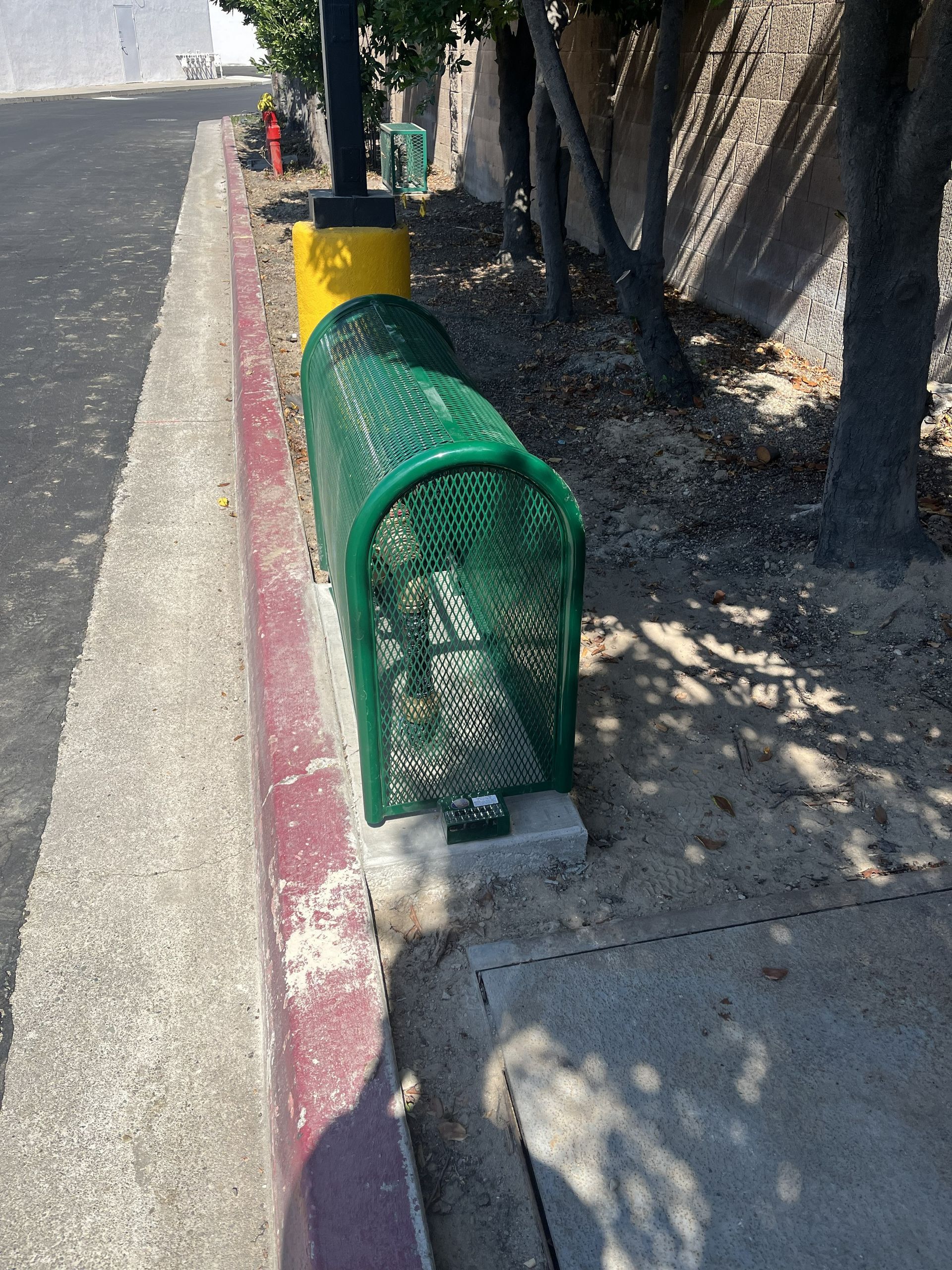 A green metal cage covering a utility box on a concrete strip next to a red-painted curb and a tree-lined path.