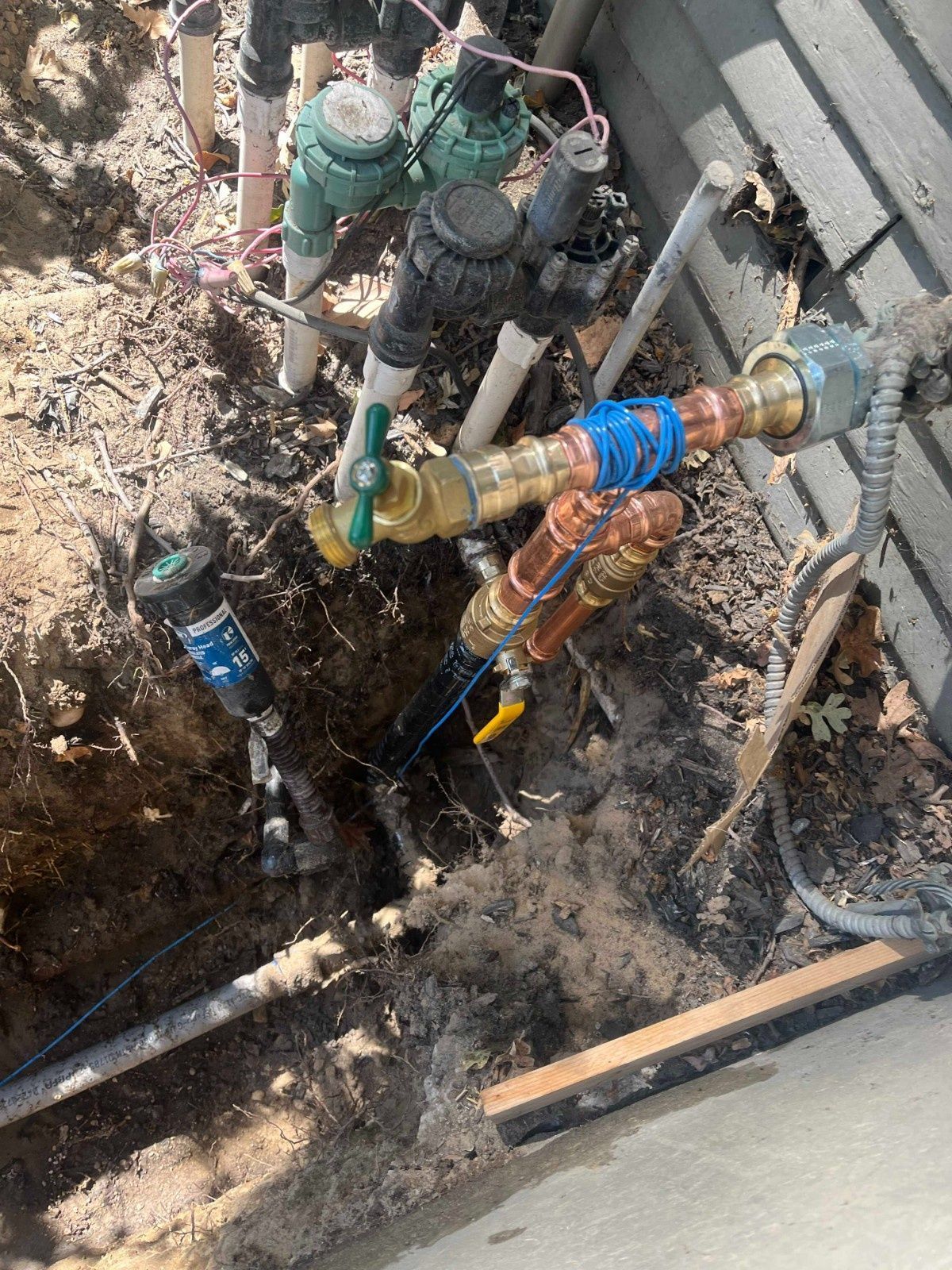 An outdoor view of an irrigation valve setup with copper pipes, a green valve handle, and wire connections in a dirt trench.