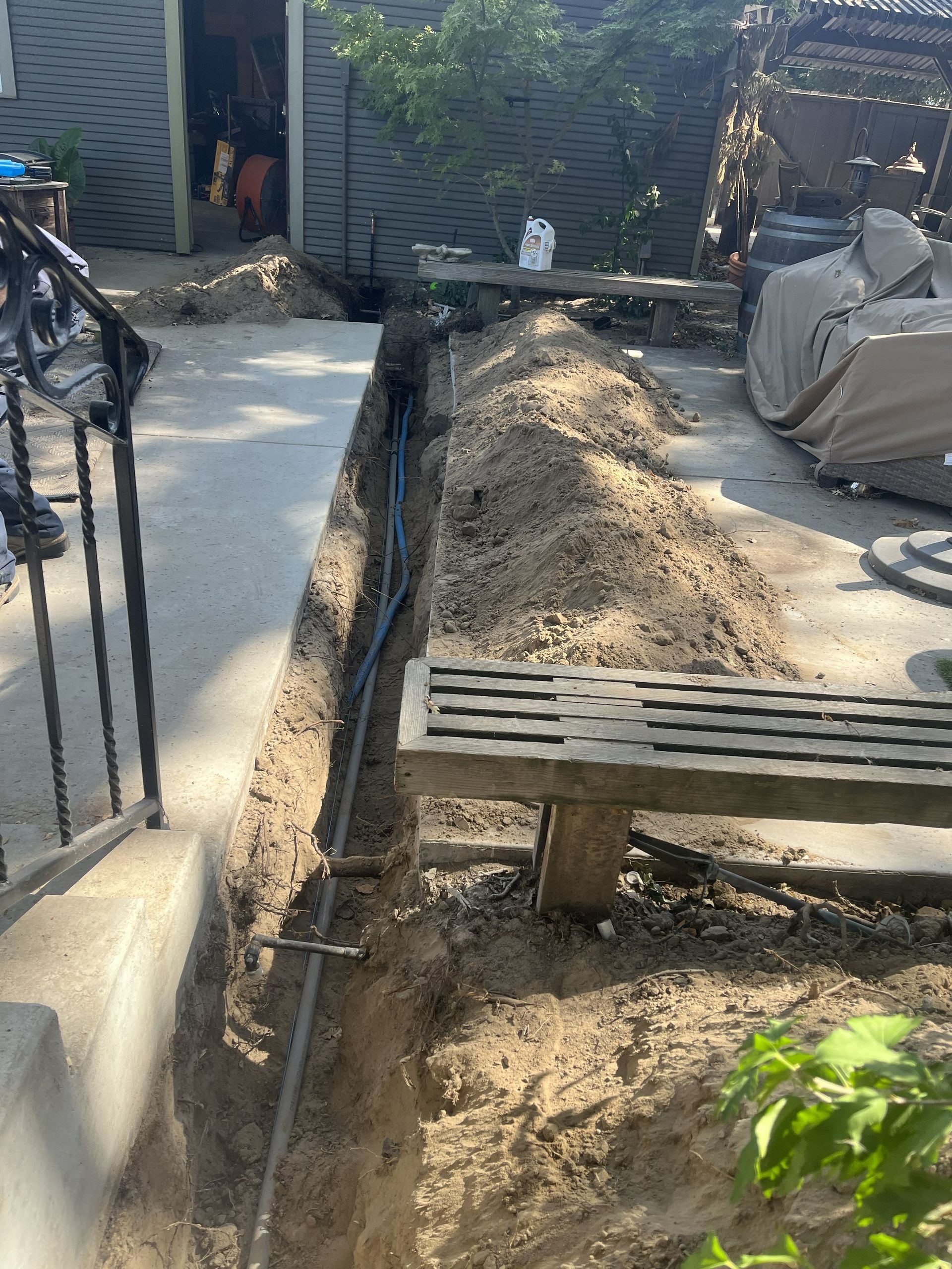A trench dug through a yard showing exposed pipes and wires leading toward a building and a covered patio area.