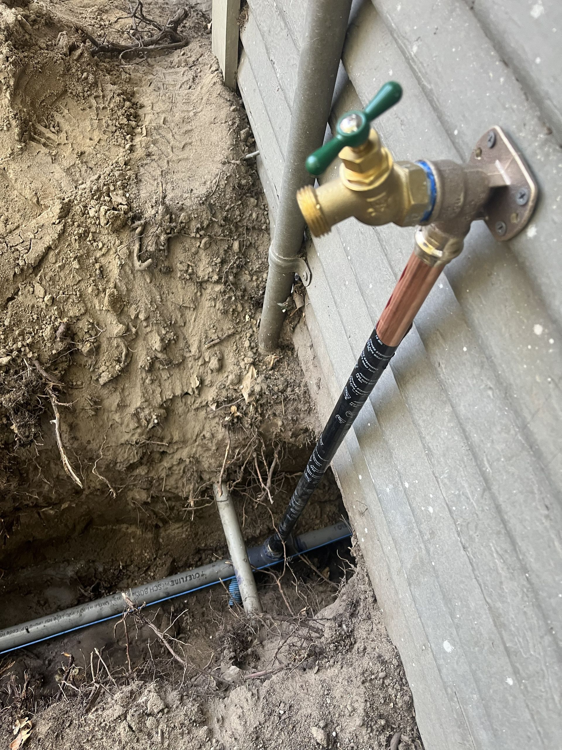 A brass outdoor faucet mounted to a siding wall, connected to a buried black pipe in an excavated dirt area.