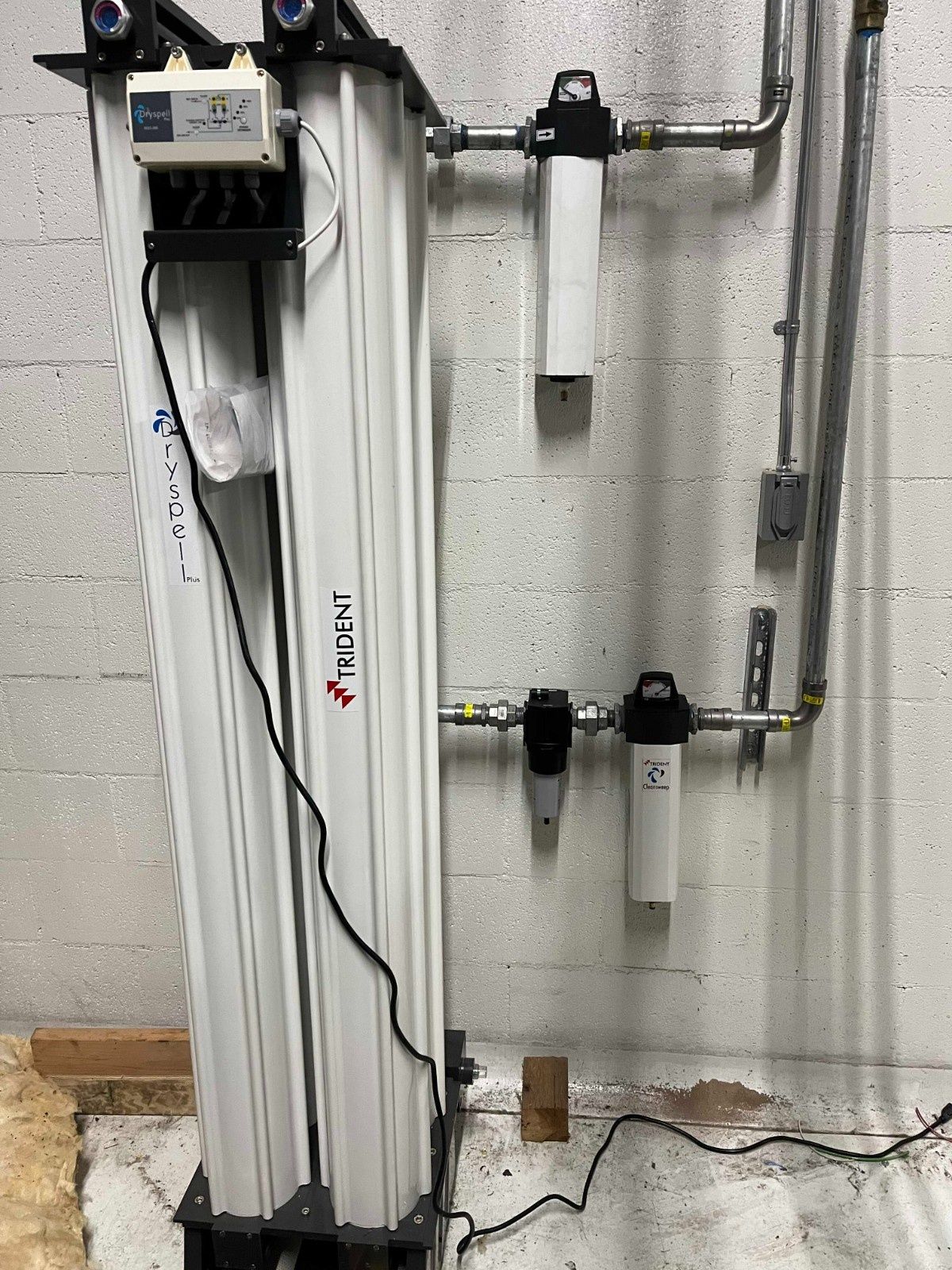 An industrial desiccant air dryer with attached filtration components mounted on a white concrete block wall.