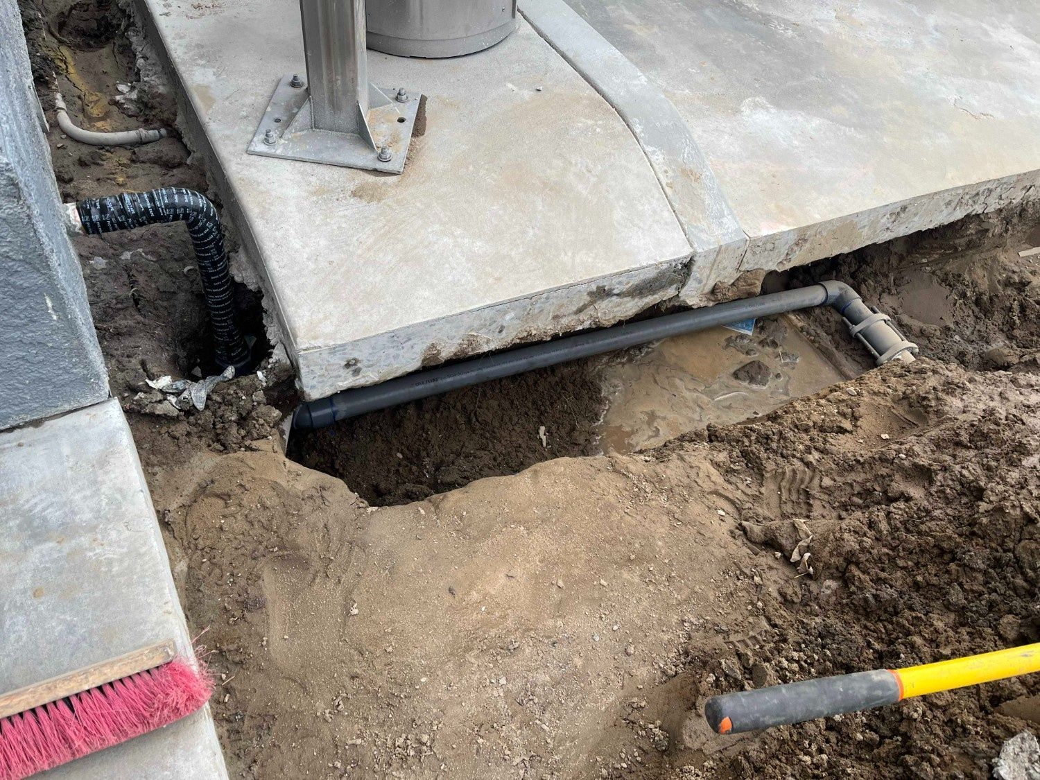 An excavated trench exposing gray pipes running along the edge of a concrete slab near a metal post.