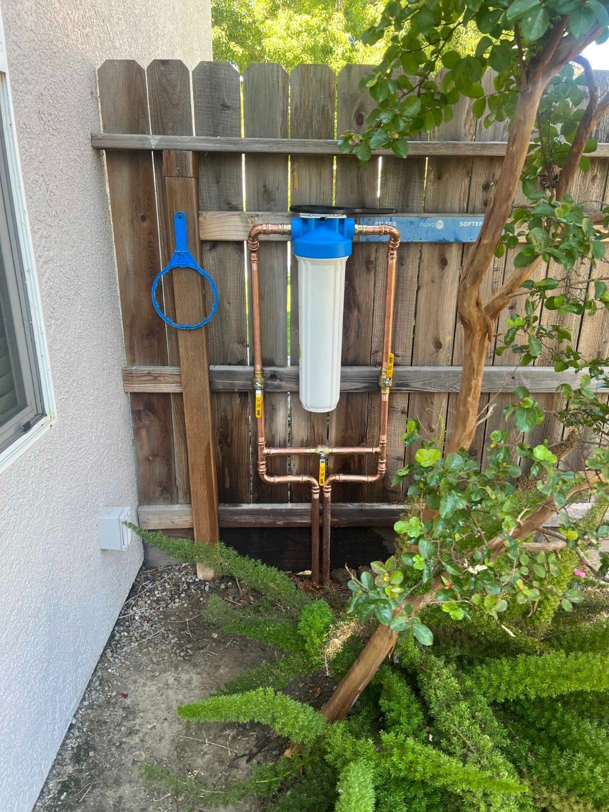 A blue and white water filter housing mounted on copper pipes against a wooden fence, with a hanging blue wrench.