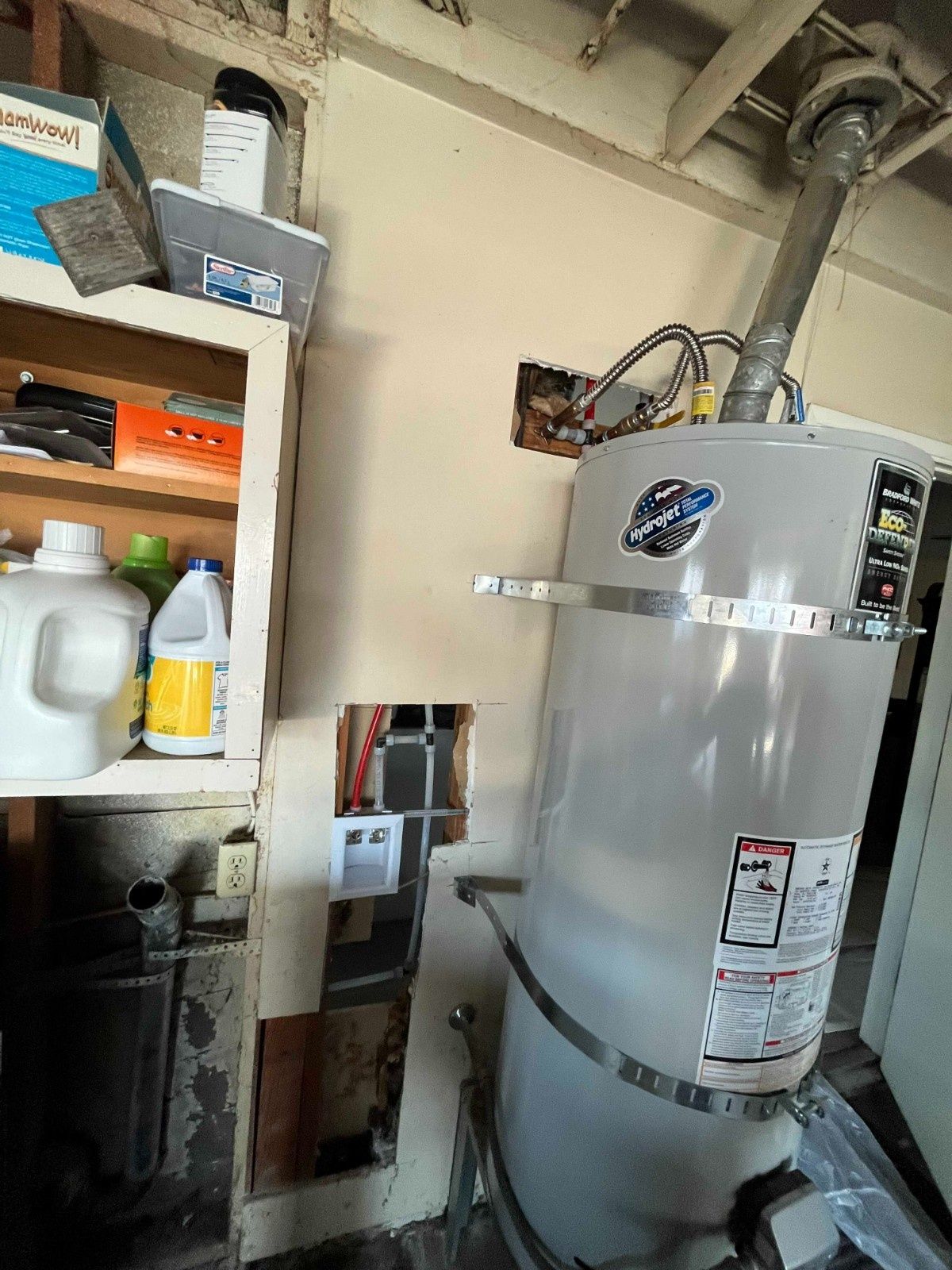 A gray water heater with metal strapping stands next to an open wall cavity showing exposed electrical wires and studs.
