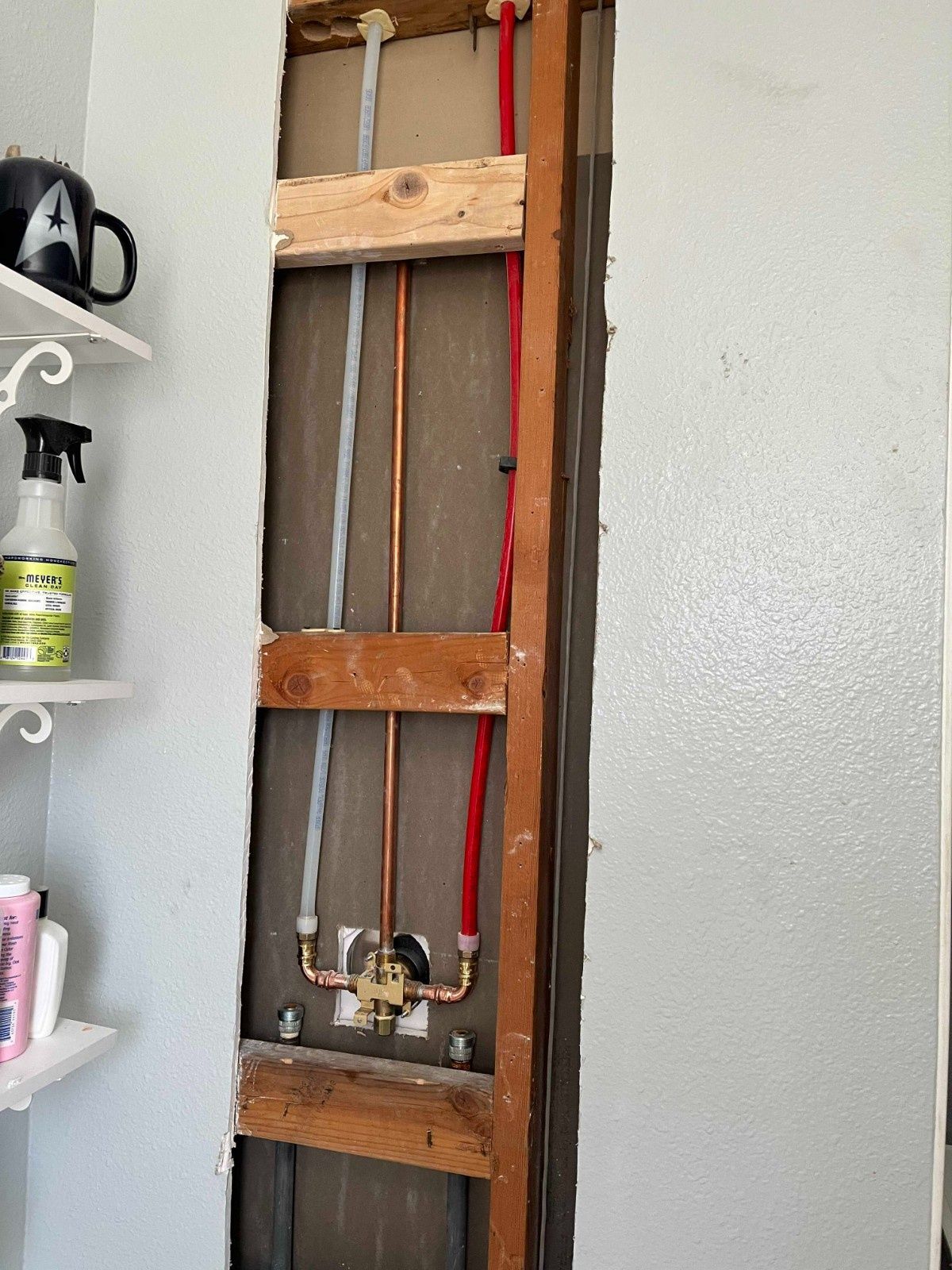 An open wall cavity showing a plumbing installation with red, grey, and copper pipes connected to a valve.