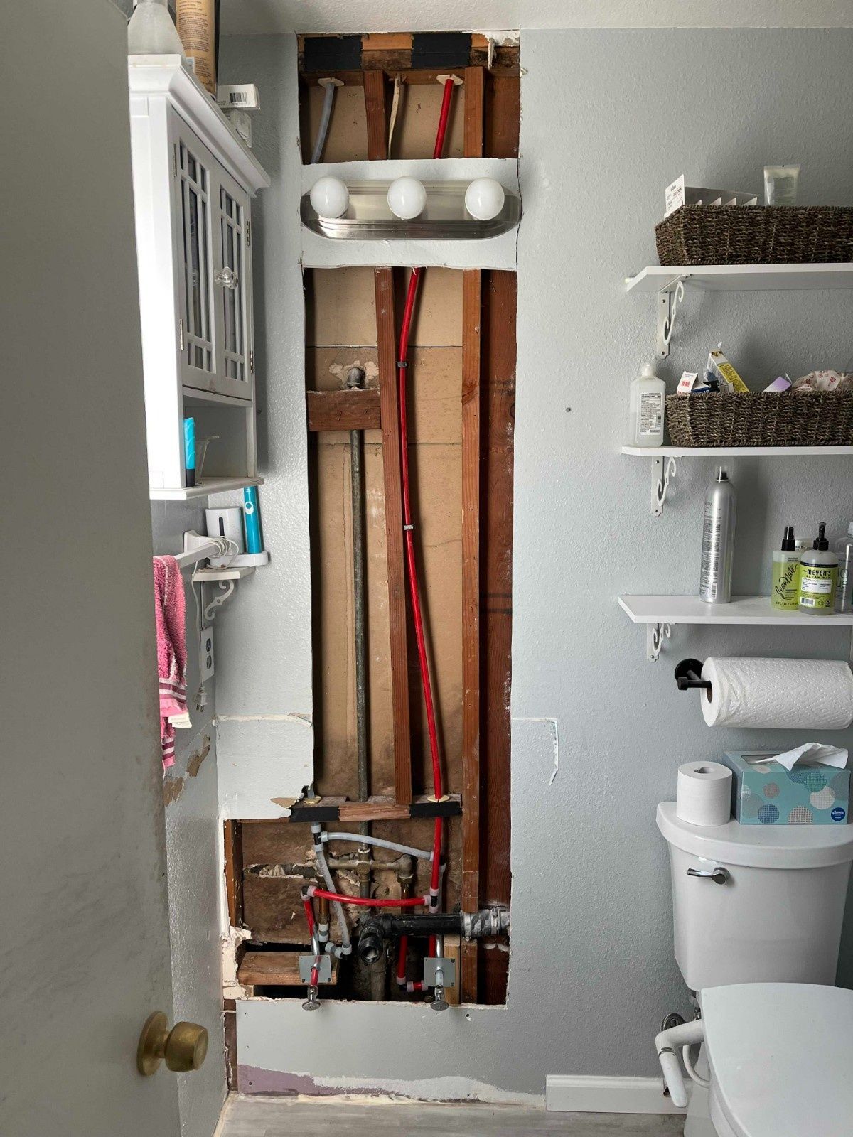 A bathroom wall with a rectangular section of drywall removed, exposing wooden studs, plumbing, and electrical wiring.