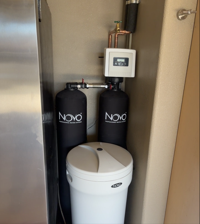 A Novo water softening and filtration system with two tall black tanks and a white salt brine tank in a utility area.