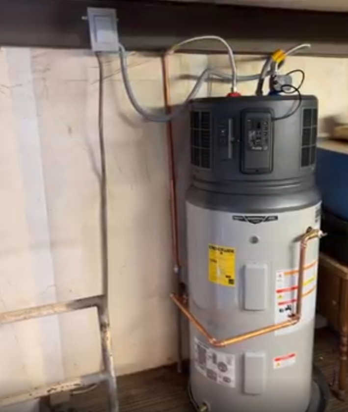 A gray hybrid electric water heater stands against a wall with electrical wiring and copper plumbing connected to the top.