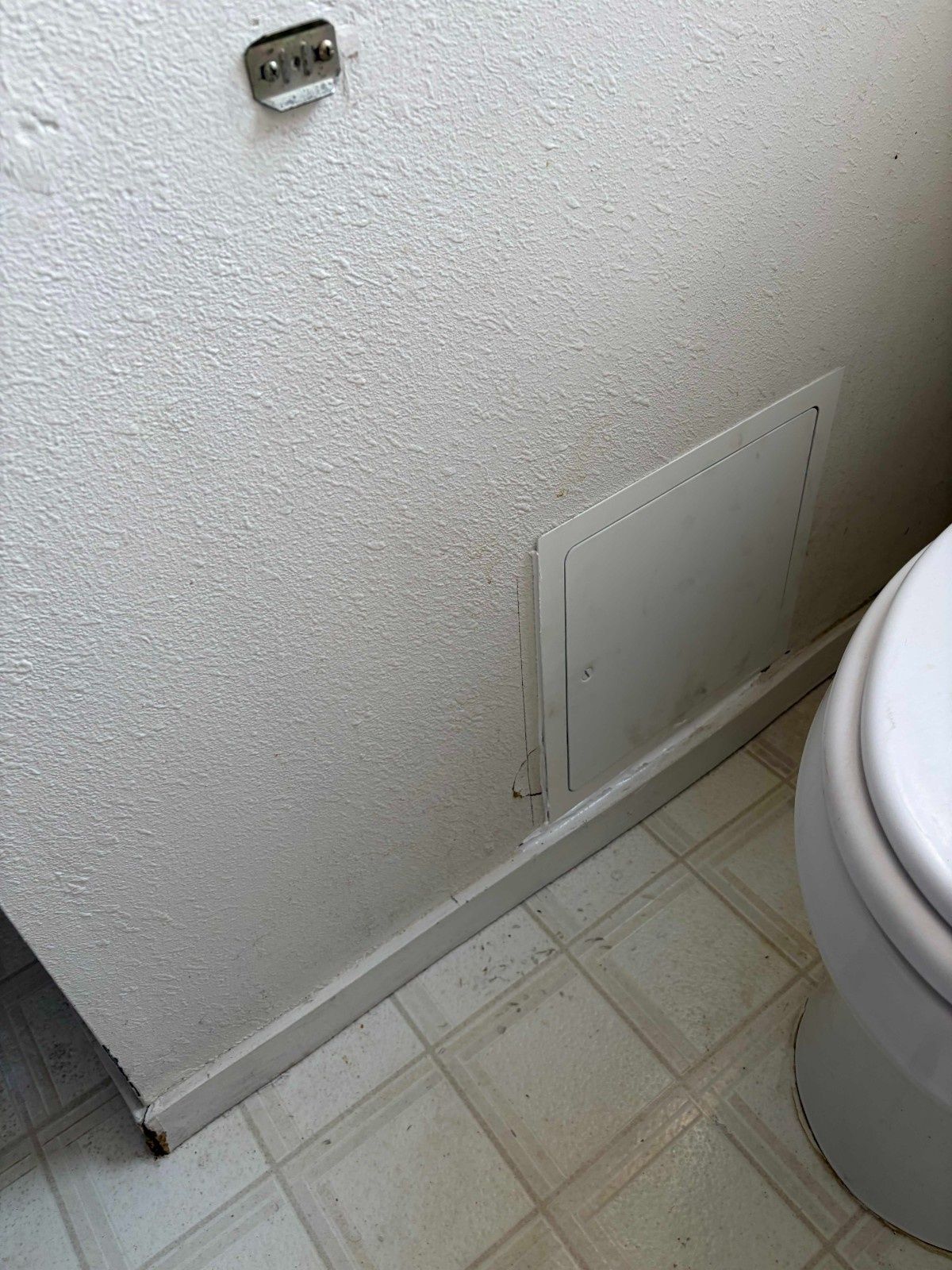 A textured white bathroom wall with a metal access panel near the baseboard and a small wall-mounted hook above.