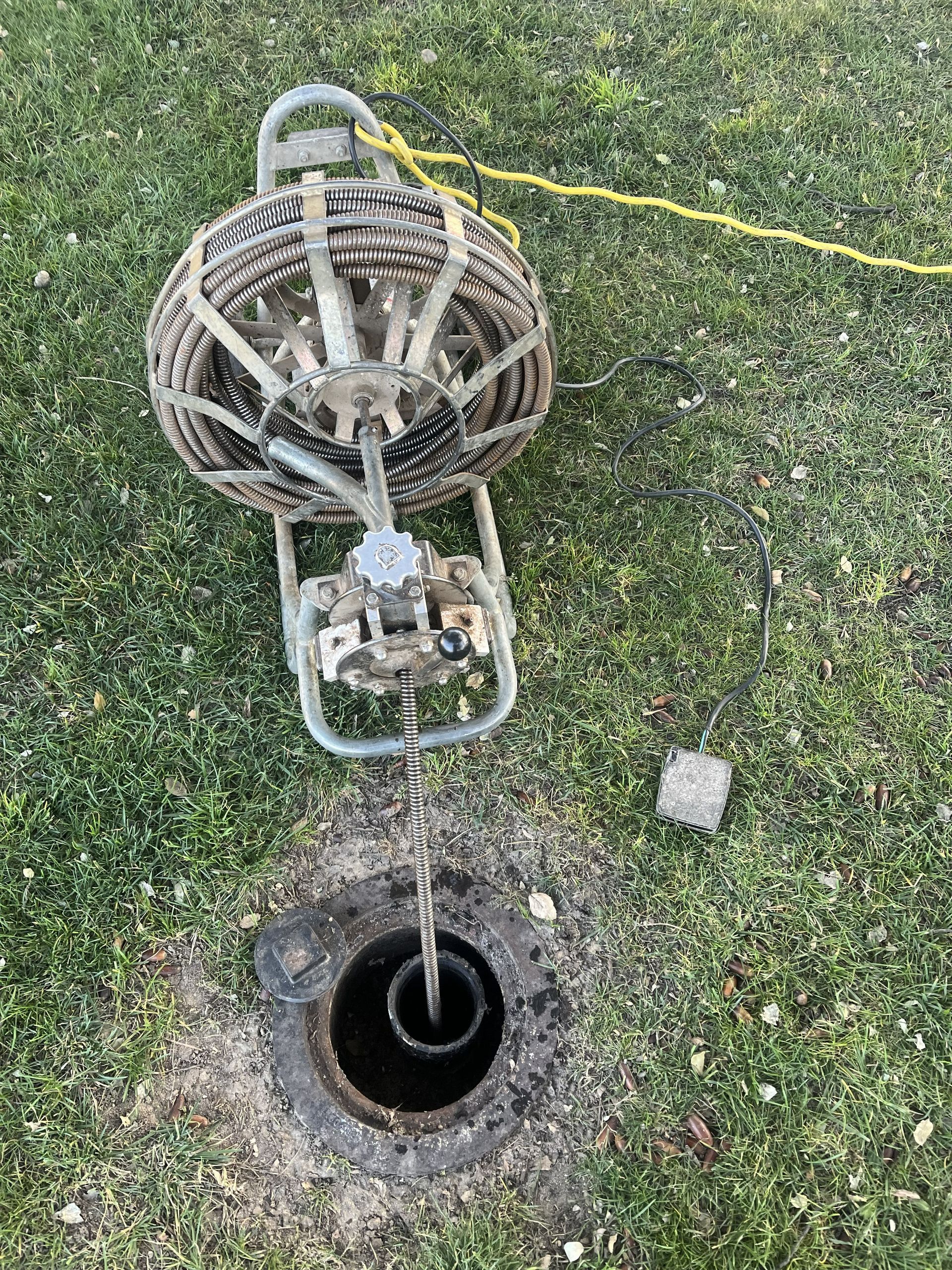 A drain cleaning tool with a coiled metal cable feeding into an open sewer access pipe in the grass.