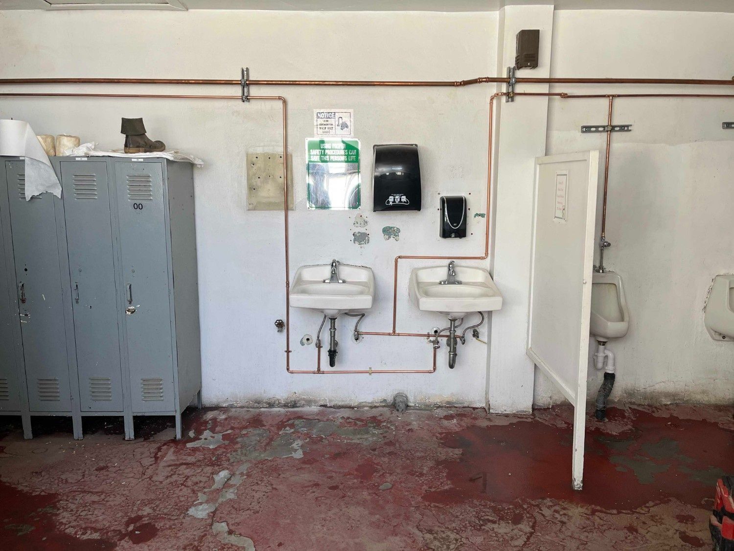 A public restroom featuring grey lockers, two wall-mounted sinks with exposed copper plumbing, and a urinal partition.