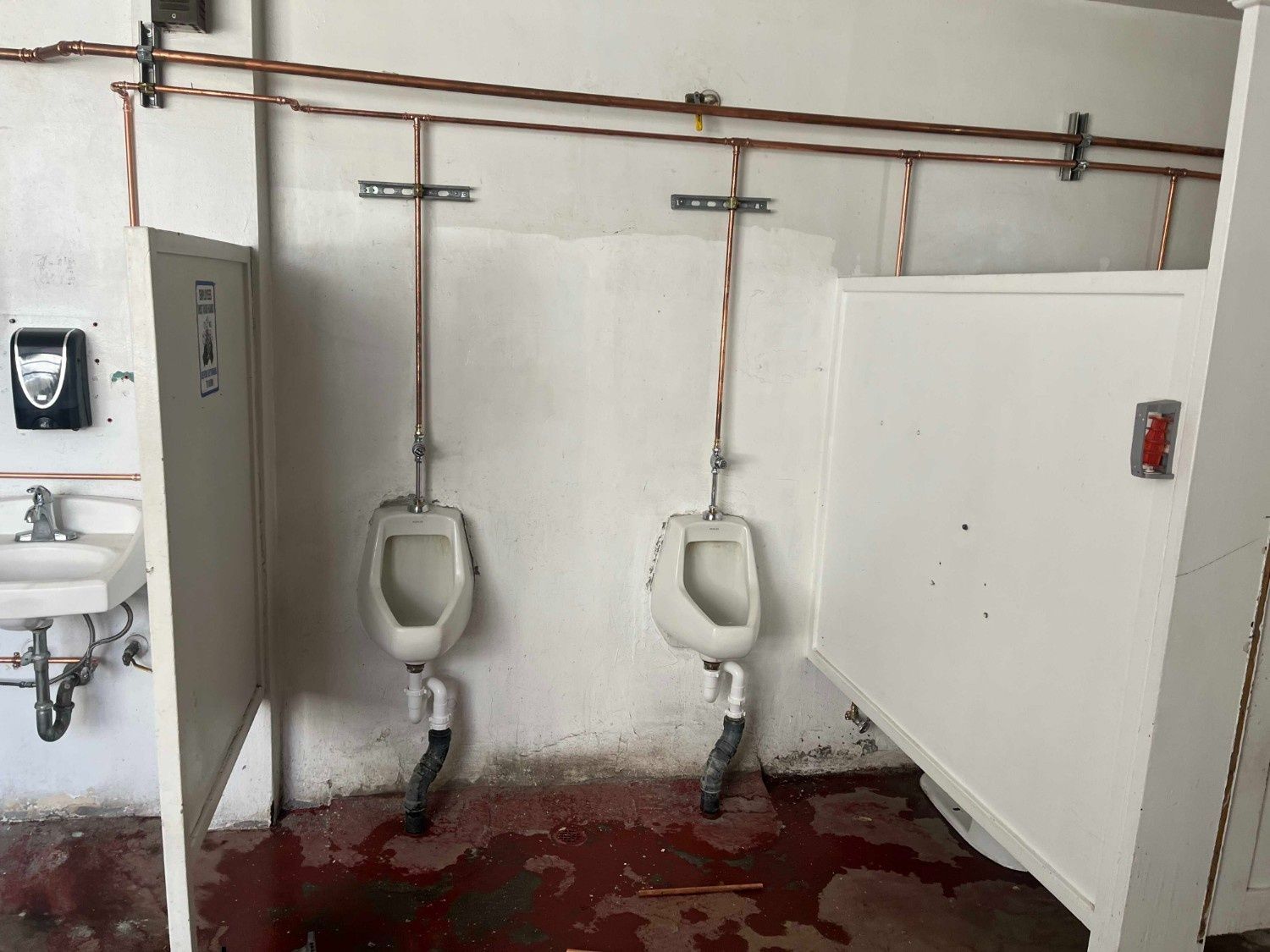 Two wall-mounted urinals in a public restroom with exposed copper piping, white stall dividers, and a stained floor.