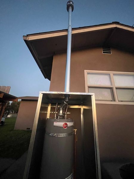 A water heater installed in an outdoor enclosure attached to a home exterior with a tall vertical metal exhaust flue.