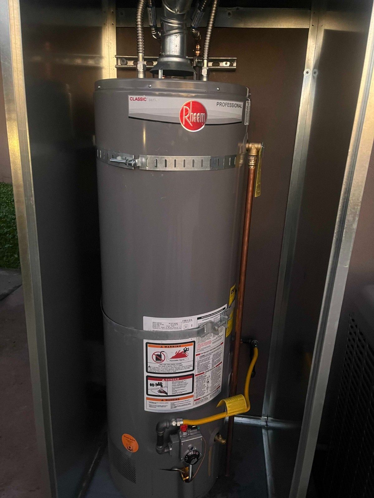 A gray Rheem gas water heater installed in an outdoor metal utility enclosure.