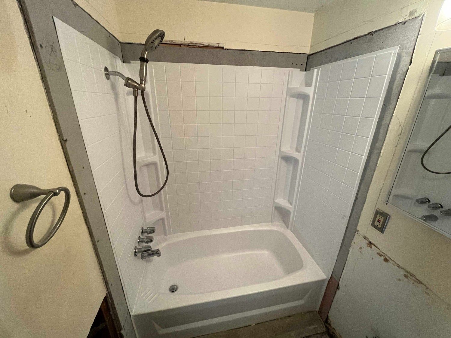 A white bathtub and shower surround with a handheld shower head, installed in a bathroom with unfinished drywall edges.