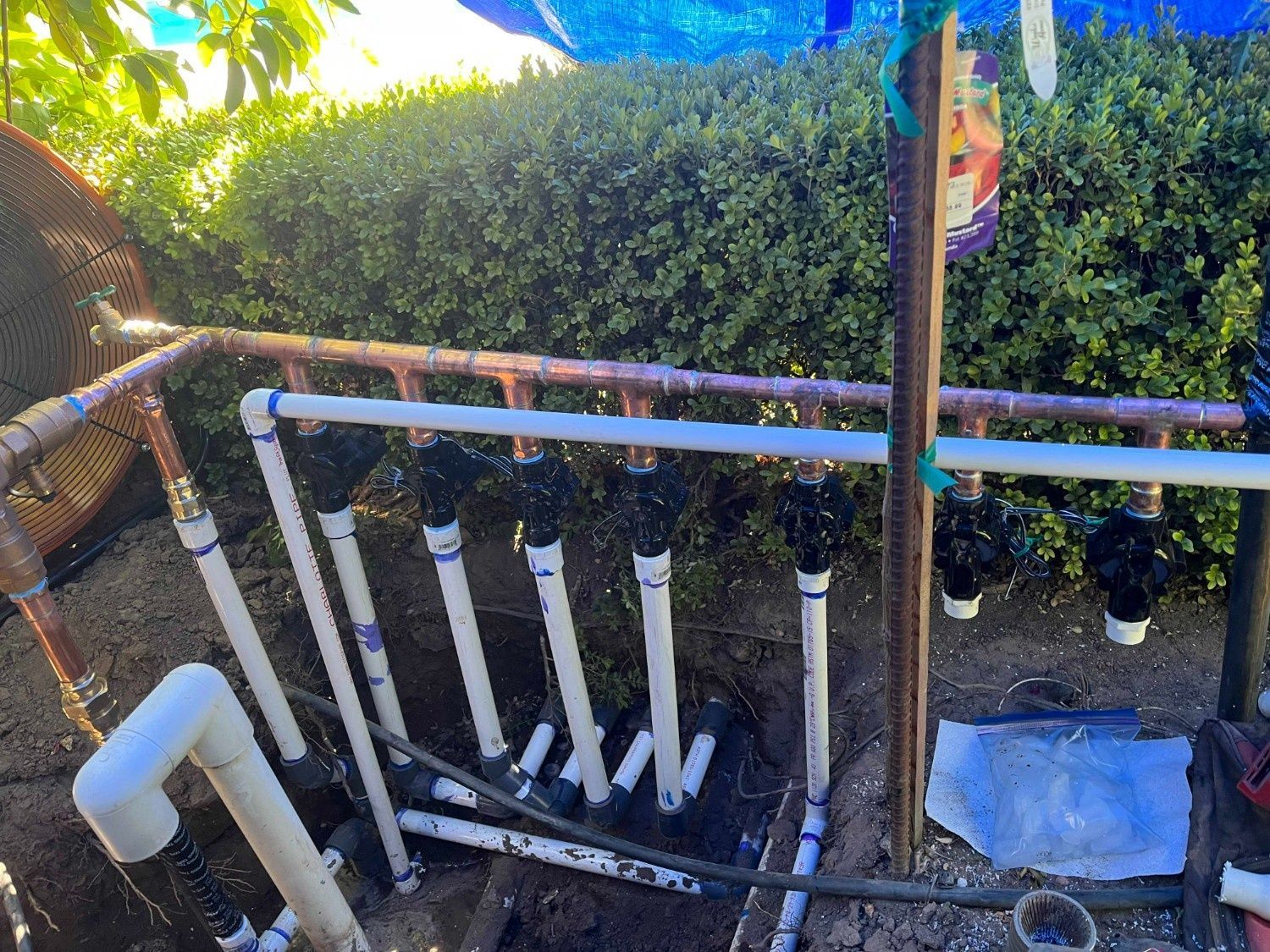 An irrigation manifold with multiple valves and white PVC pipes installed outdoors in front of a hedge.