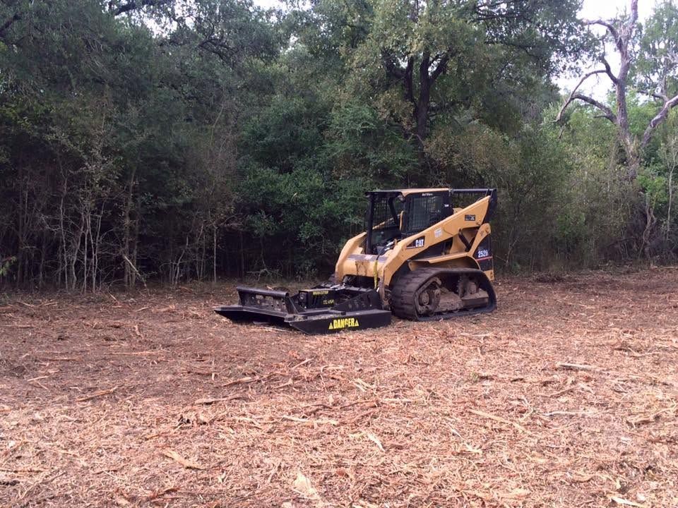 Yellow skid steer with brush cutter on a dirt clearing beside dense trees.