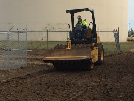 Yellow bulldozer grading dirt near a chain-link fence beside a large beige building