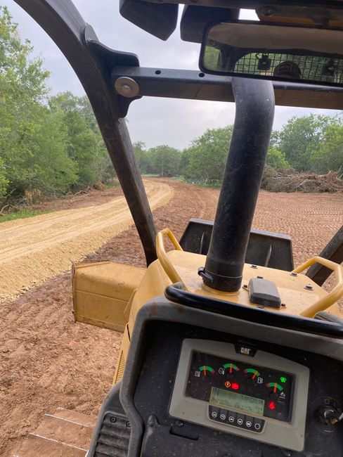 View from a tractor cab driving on a dirt road beside trees, with the dashboard visible.
