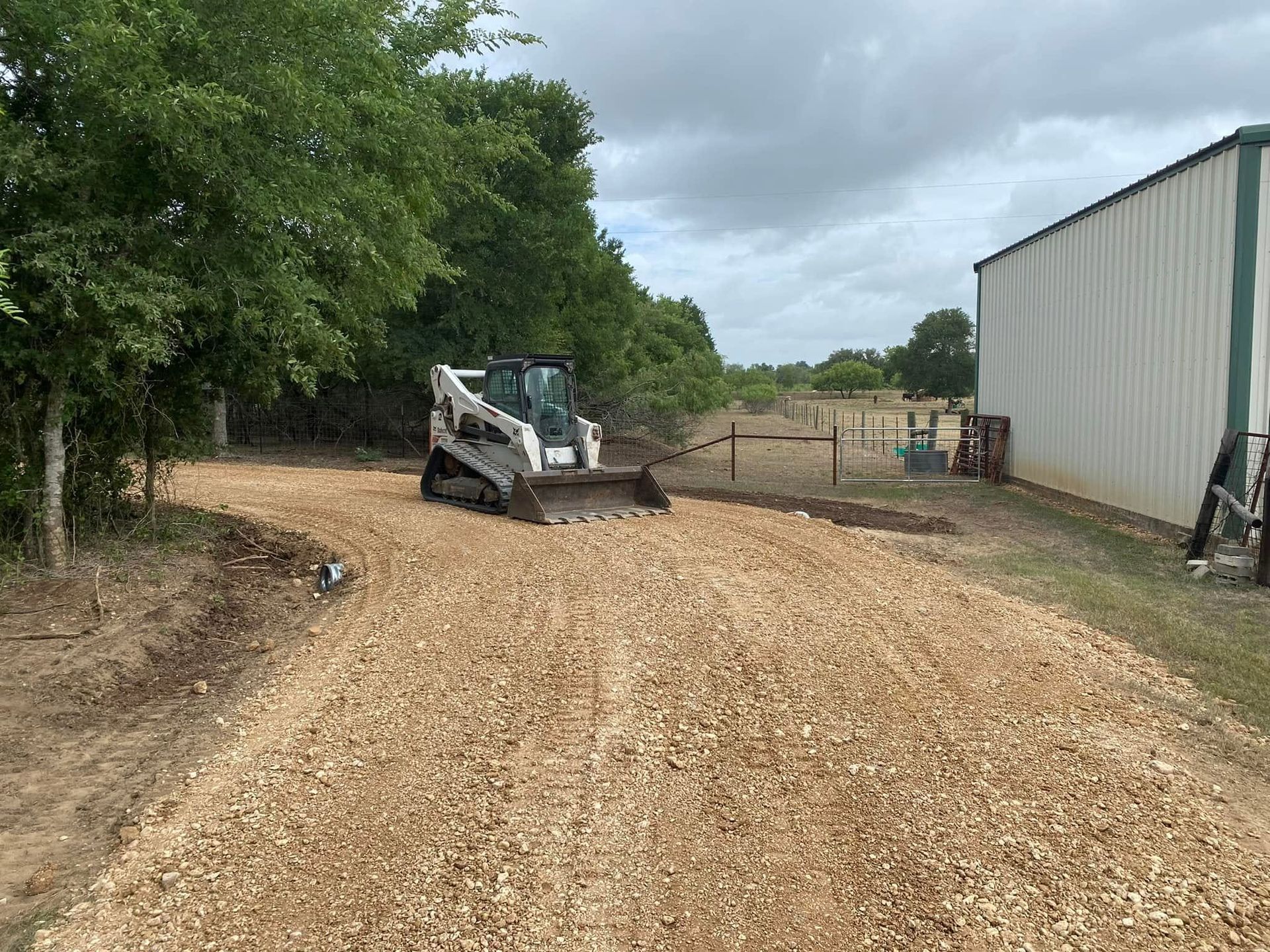 Skid steer on a gravel drive beside a shed and trees under cloudy skies