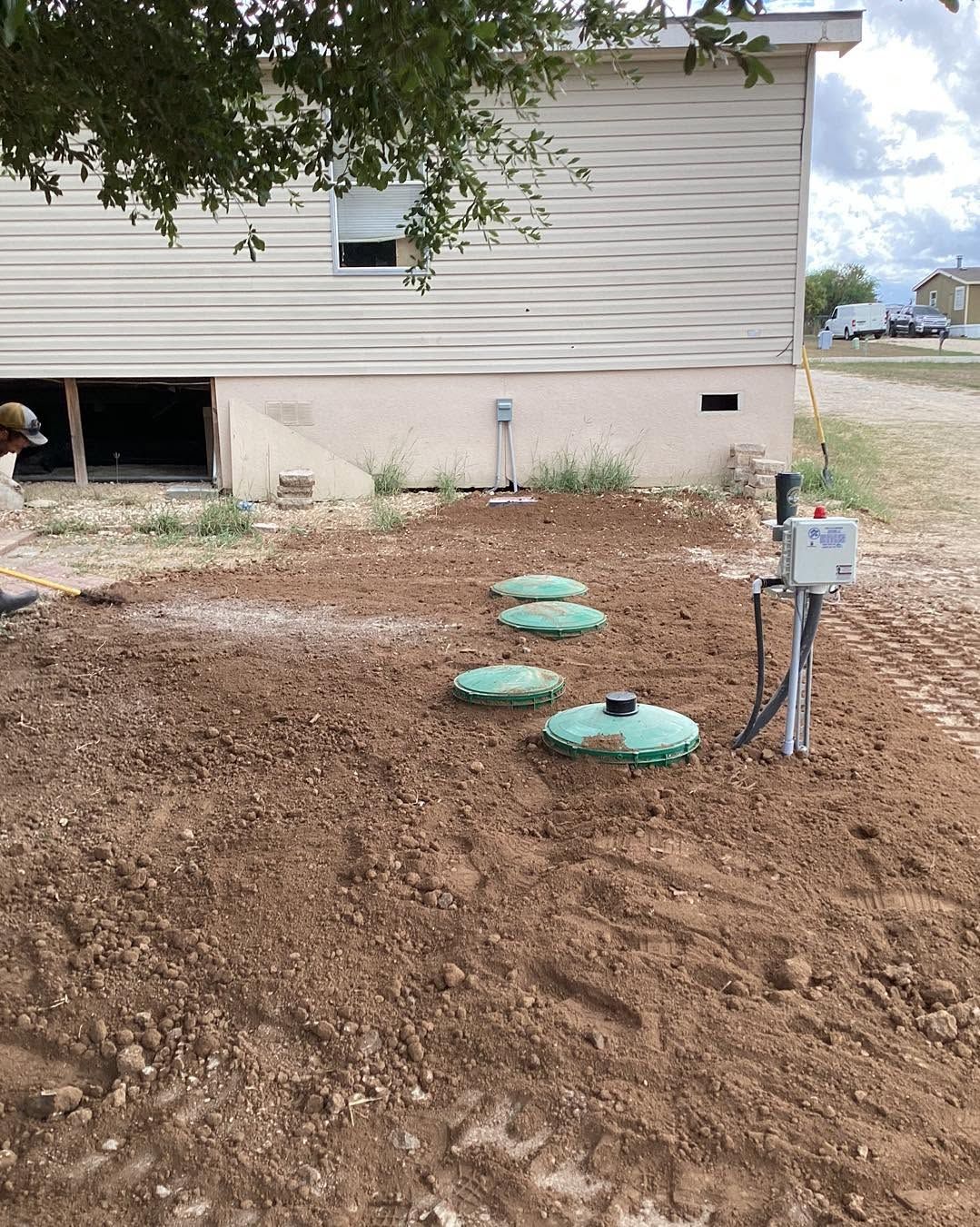 Mulched yard with three green utility lids beside a house and meter box under a tree