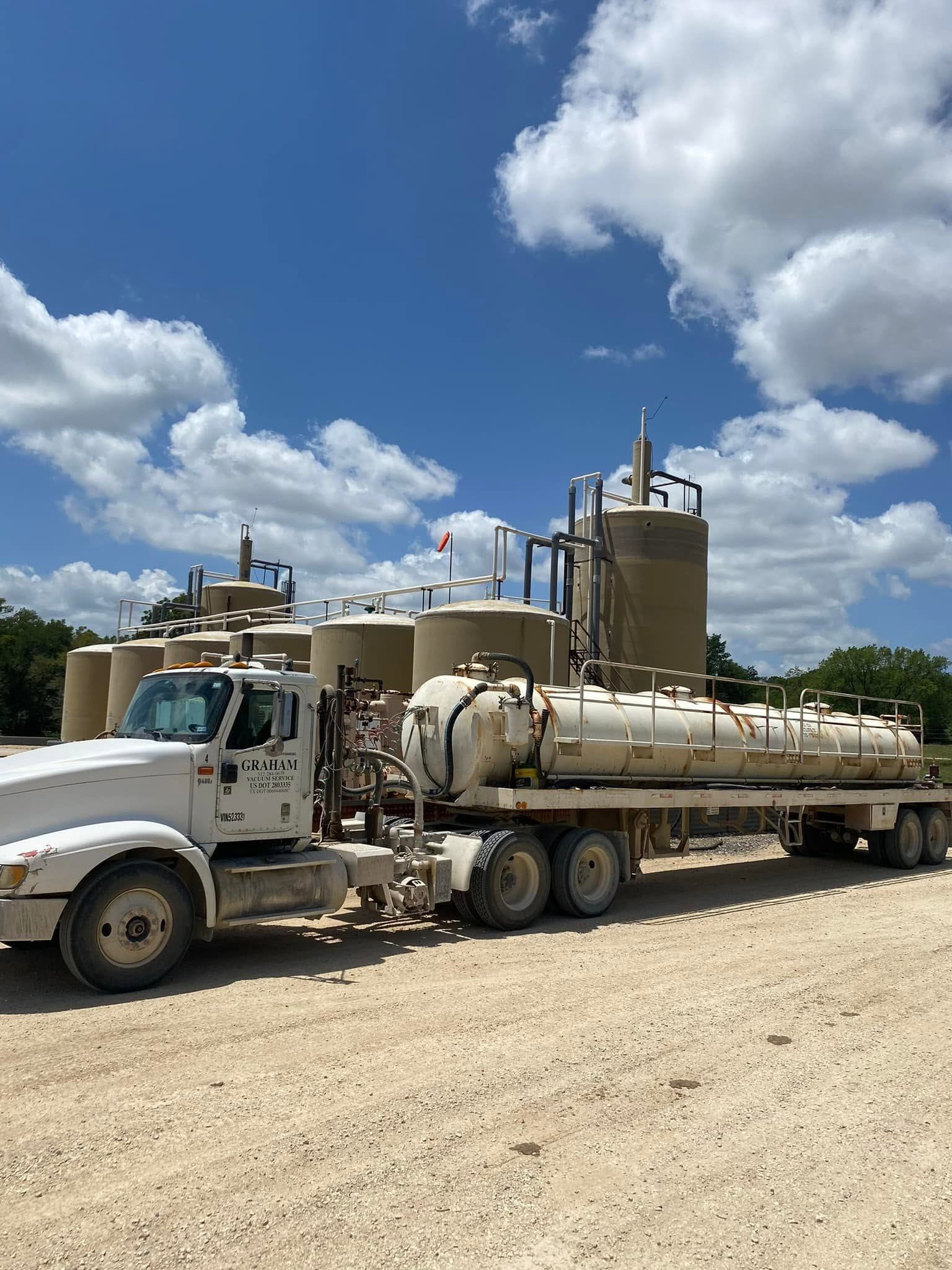 White tanker truck on a gravel road beside industrial tanks under a bright blue sky with clouds