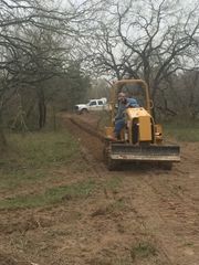 Yellow bulldozer clearing a dirt path in a wooded area, with a white pickup truck nearby.