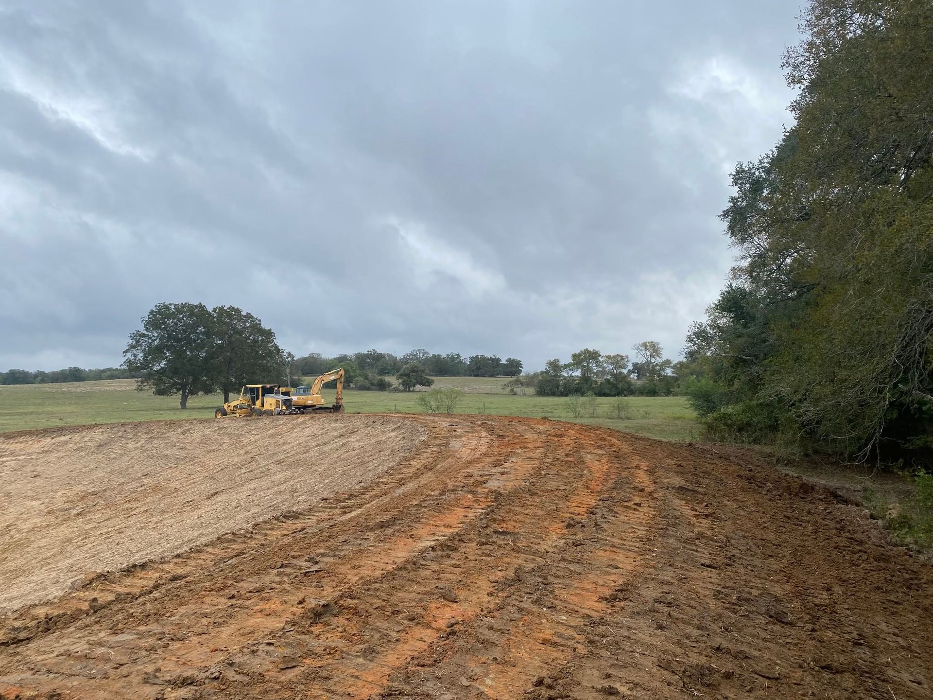 Construction machinery grading a dirt field under a cloudy sky, with trees and open land nearby.